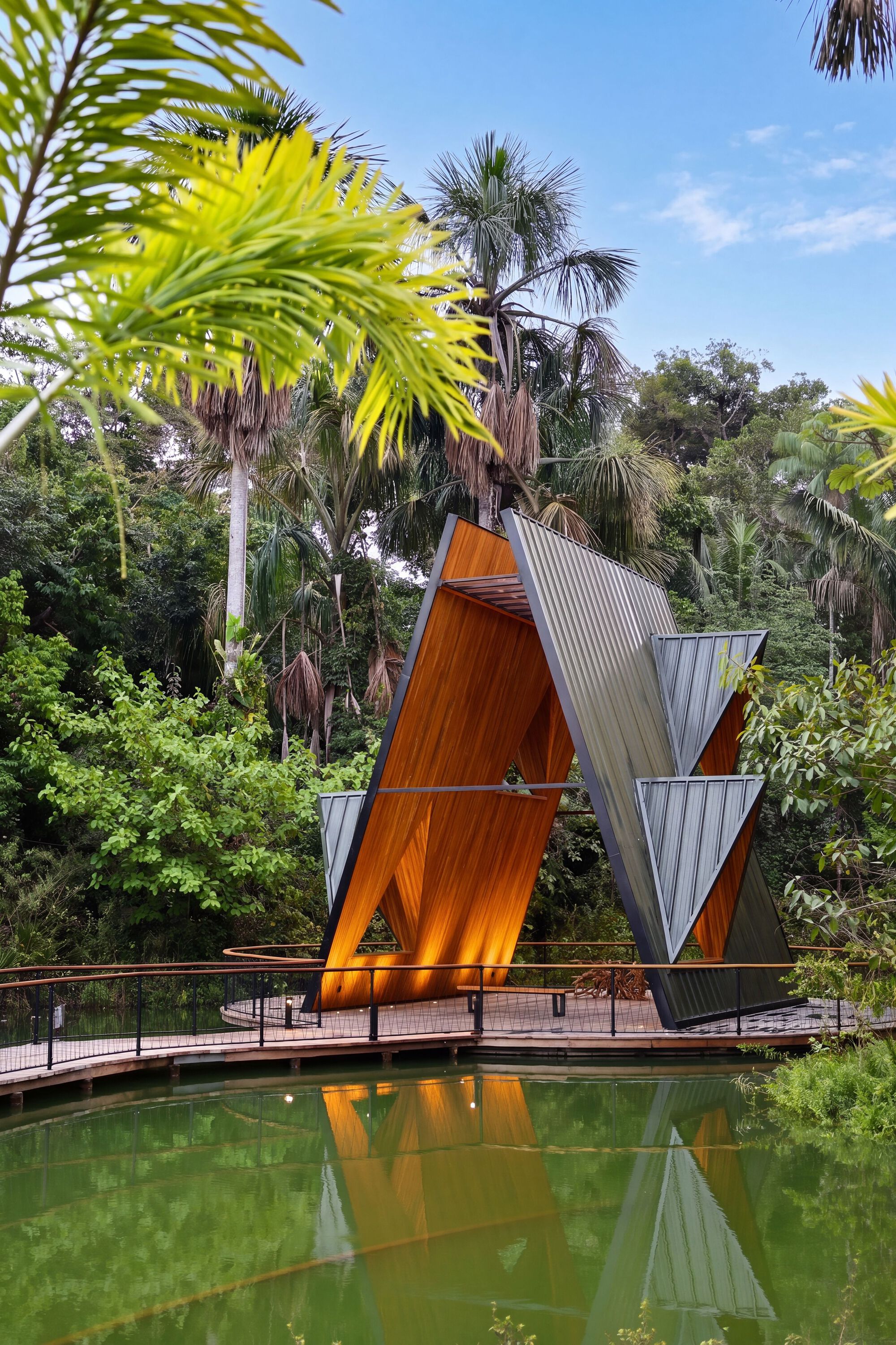 Warm timber-lined interior of the V-shaped pavilion glowing at dusk, mirrored in the green pond beneath palms and blue sky
