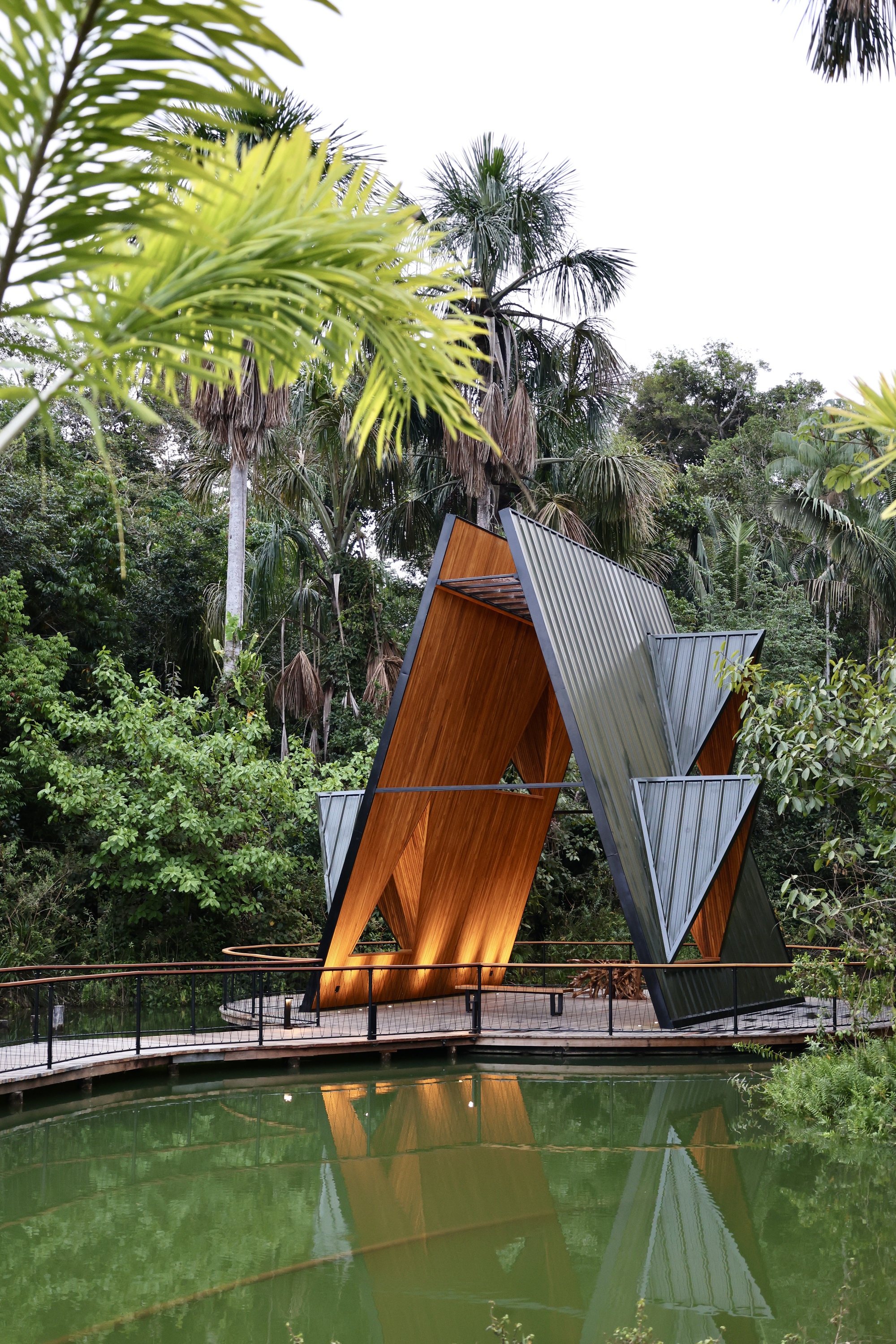 Folded timber and metal pavilion on a pond deck, its glowing wood interior reflected in green water among palms