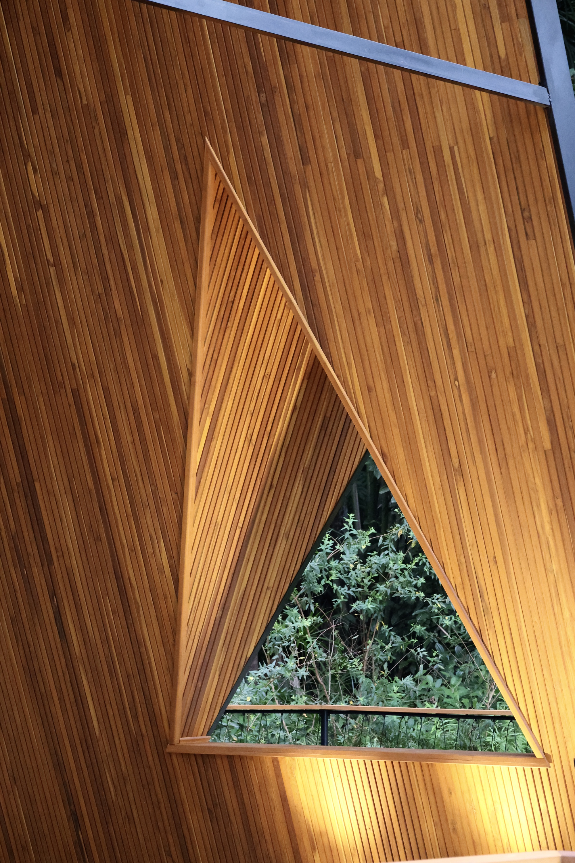 Triangular window cut into slatted timber wall, framing dense green foliage and a thin railing beyond