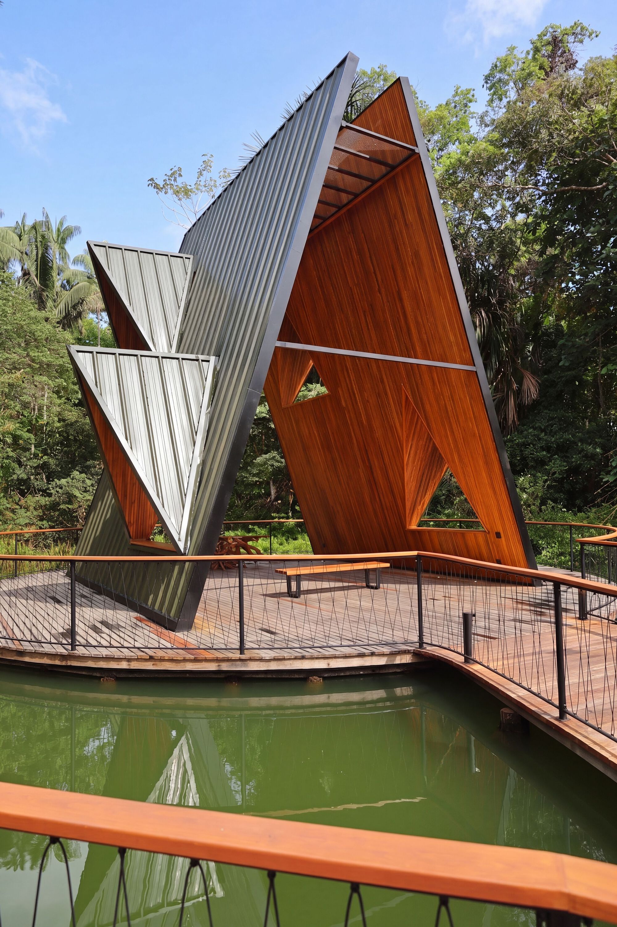 Angled pavilion with metal-clad and timber faces on a pond-edge deck, a bench beneath and sunlit forest behind