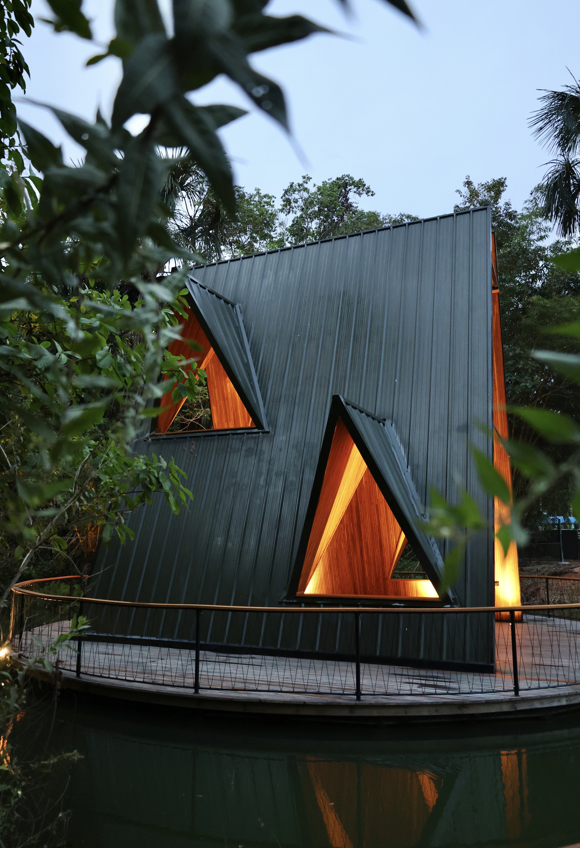 Dark standing-seam metal facade with two triangular glowing timber-lined windows above a railed deck, seen through leaves at dusk