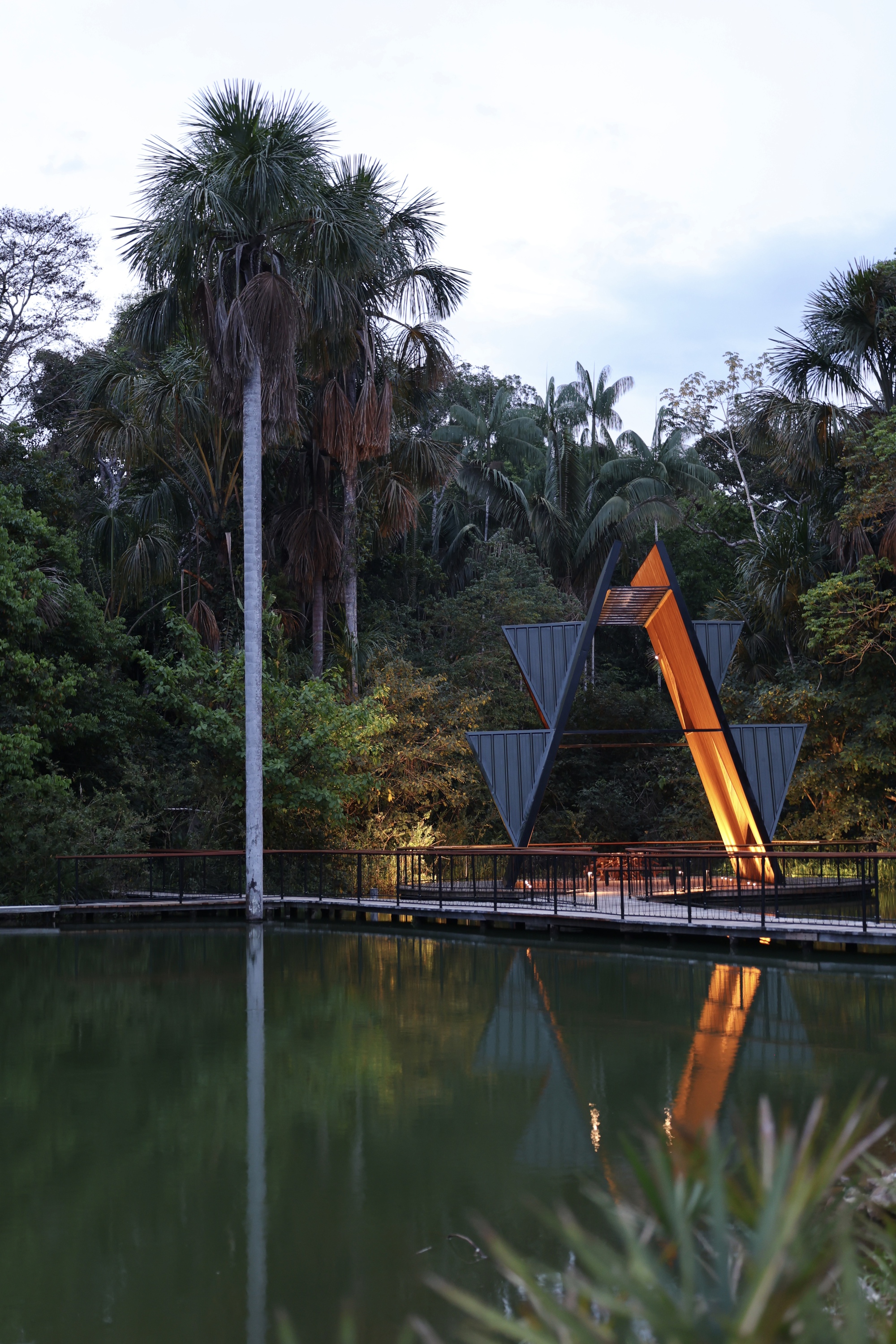 Open triangular frame of the pavilion with an amber lit timber edge, reflected in a still pond at dusk