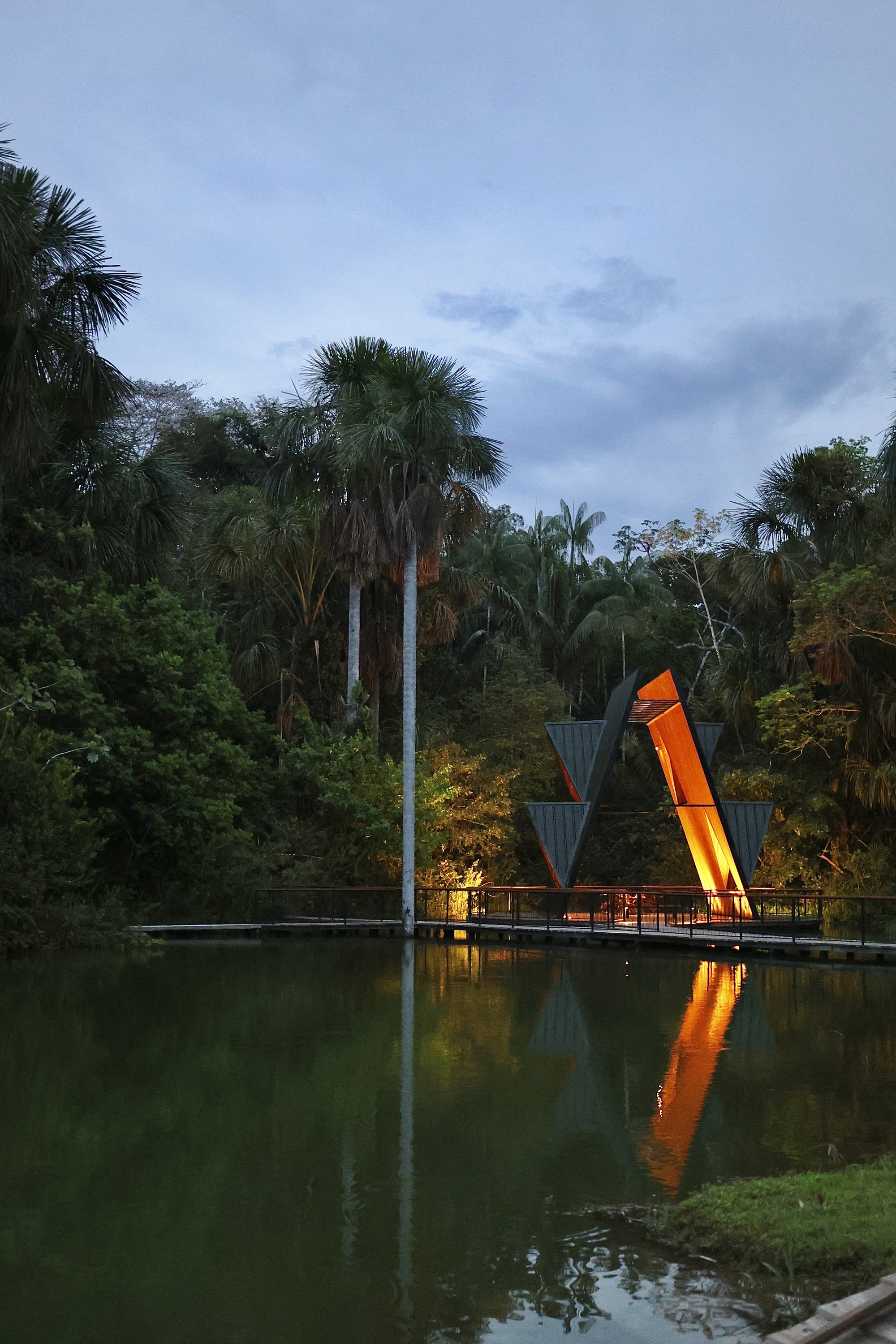 Distant view of the A-shaped pavilion with a glowing orange interior edge, mirrored in dark pond water under cloudy twilight