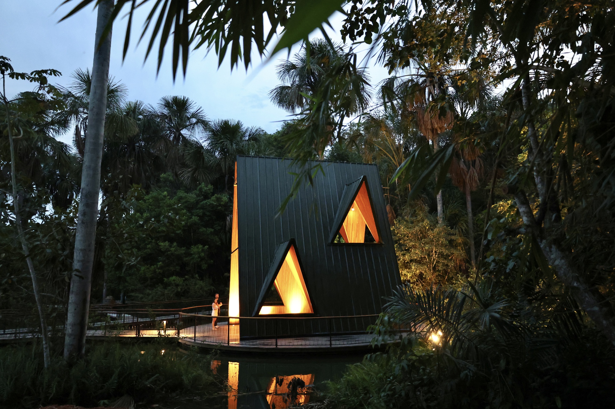 Black metal clad pavilion with two triangular windows glowing amber, a person standing on the curved deck among palms at dusk