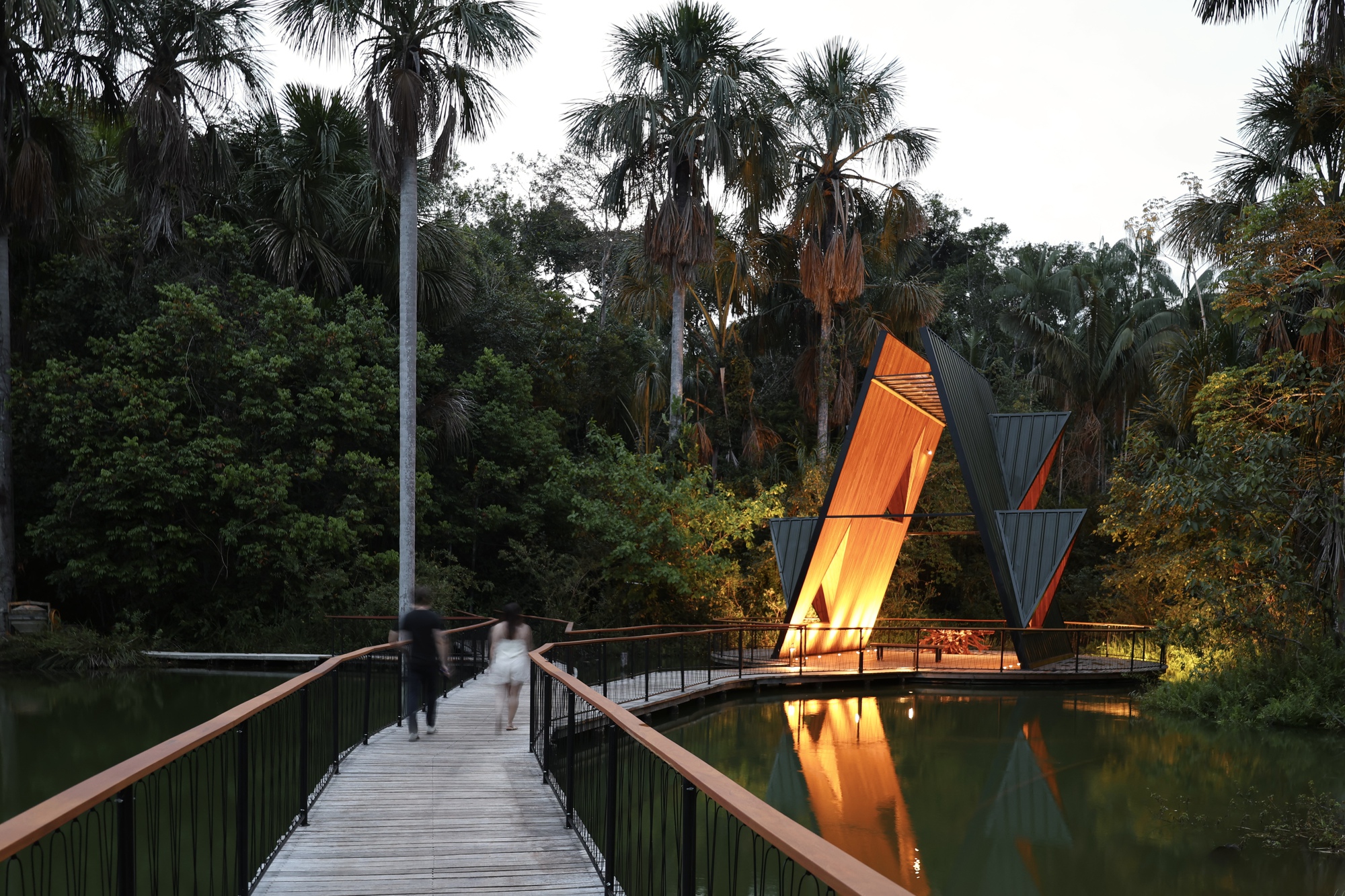 Two people walking along a timber boardwalk with rust-colored handrails toward the glowing angular pavilion in the evening forest