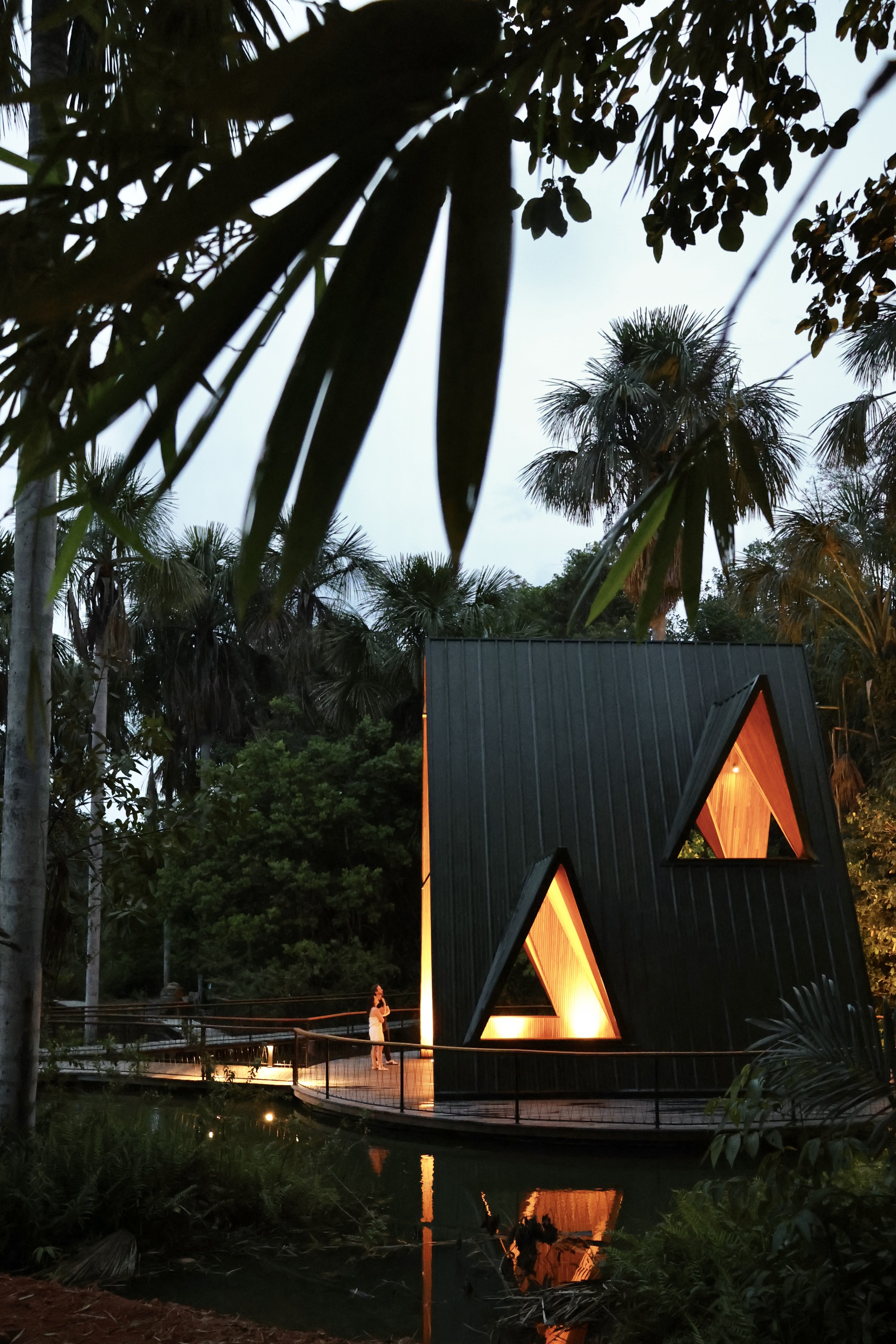 Black triangular pavilion with lit timber-lined window openings, a person on the deck and palm fronds hanging in the foreground at dusk