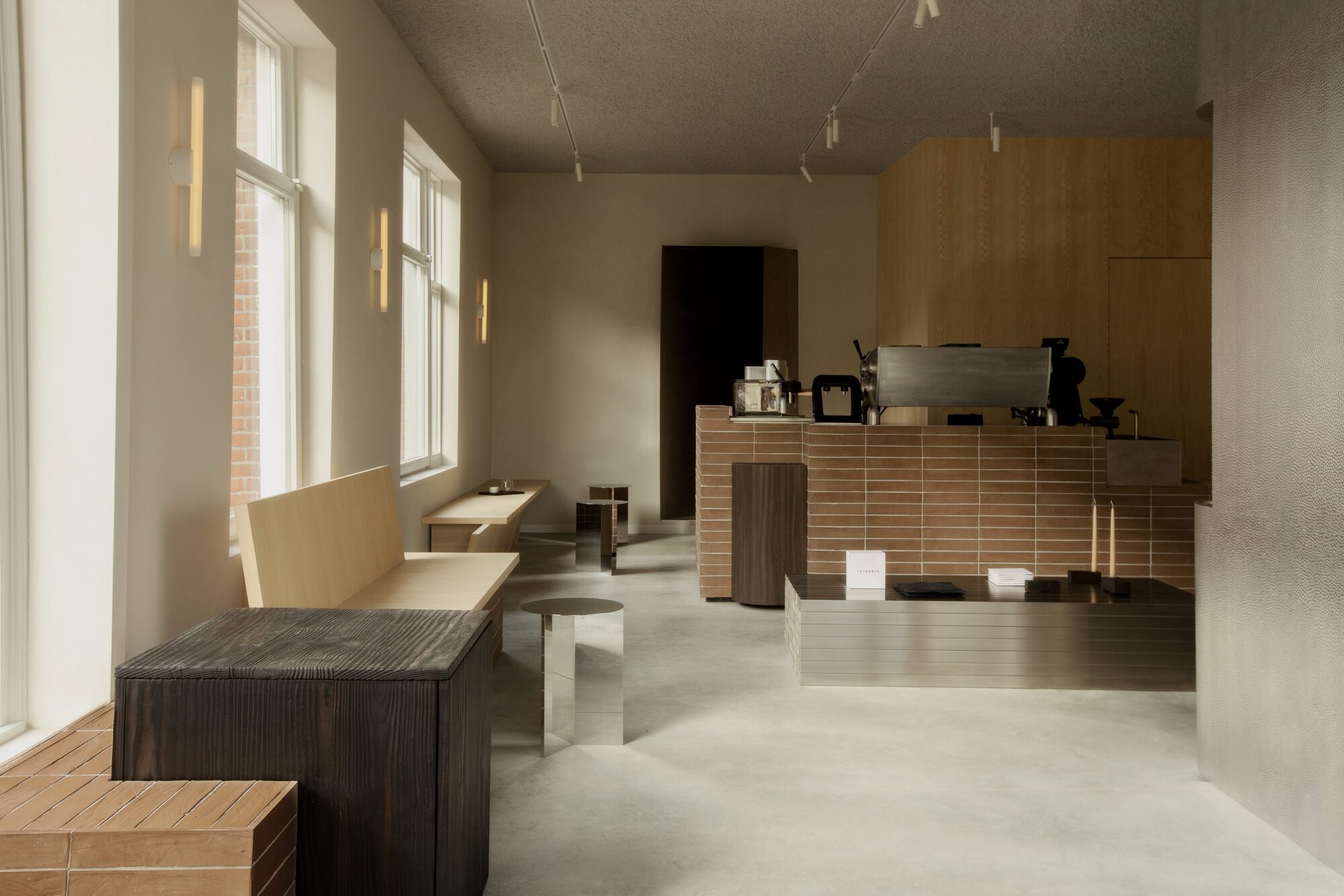 Coffee bar with a brick-tiled counter, steel-clad table and charred timber bench, lit by daylight from tall windows