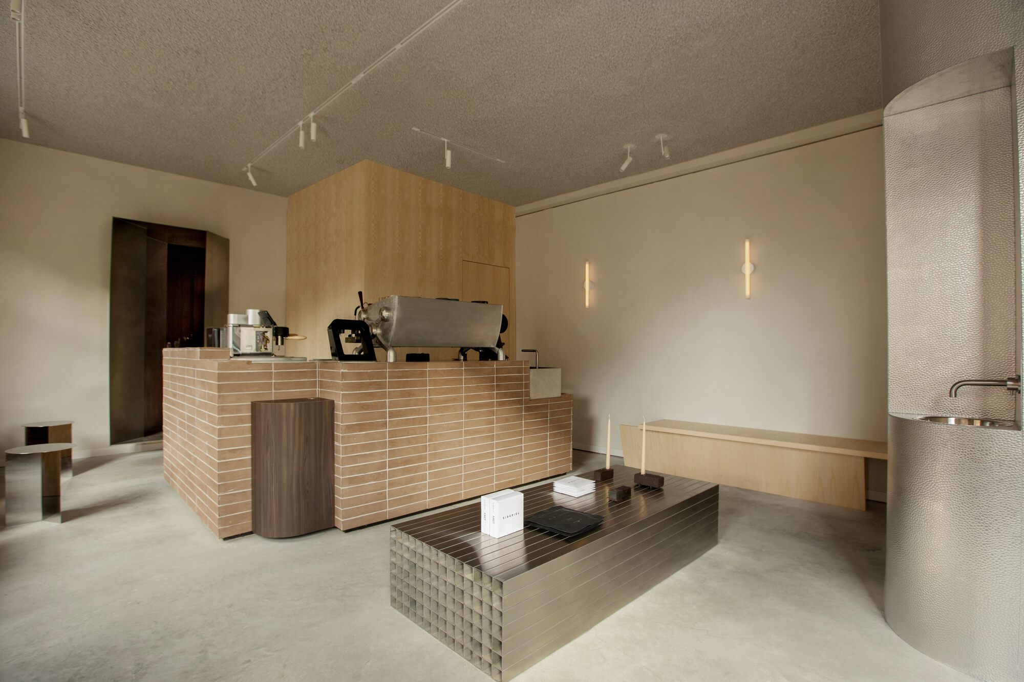 Brick-tiled service counter beside a ribbed steel table and a hammered metal sink column under a textured ceiling