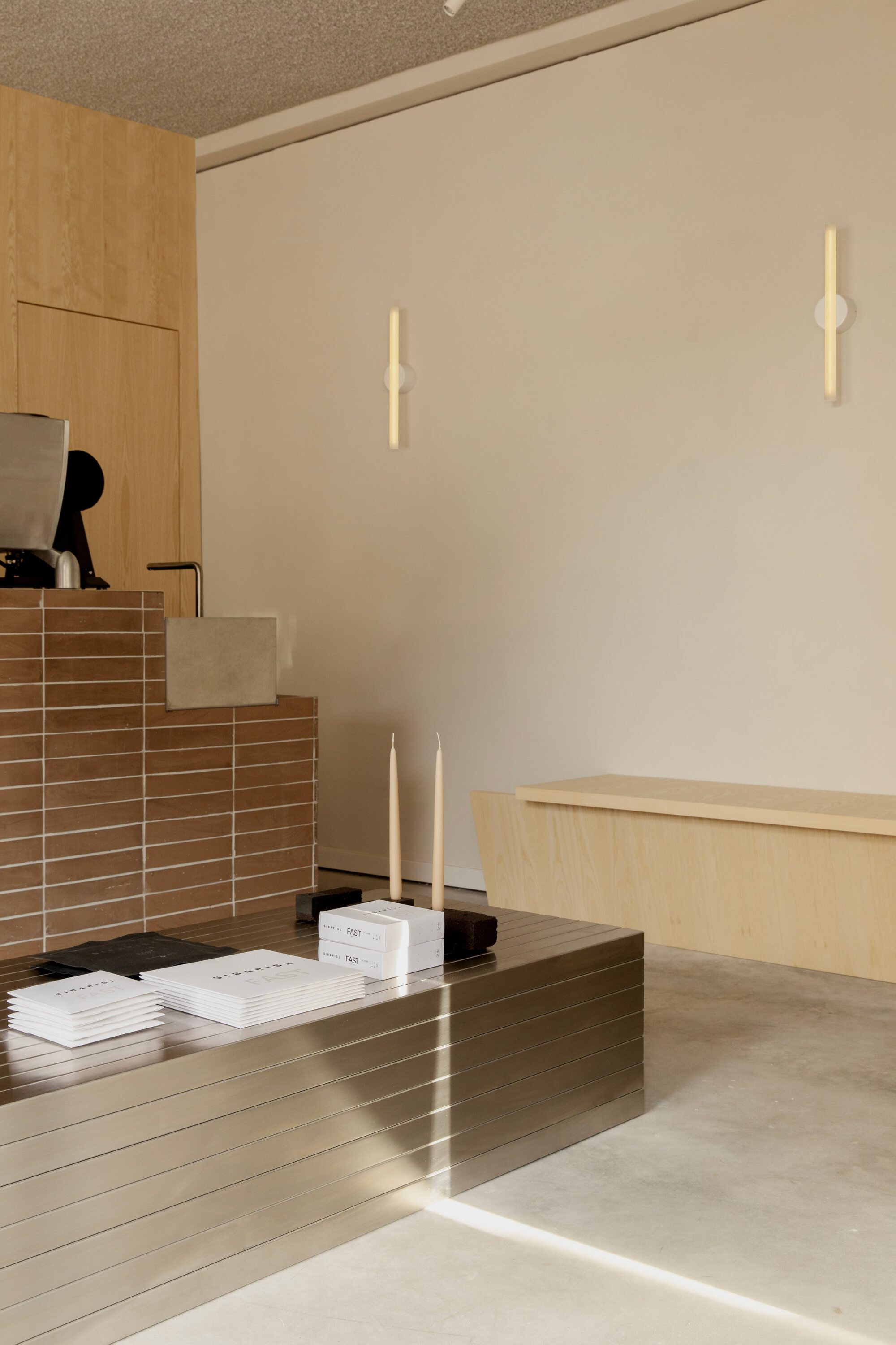 Ribbed stainless steel table holding books and candles in front of a pale plywood bench and slim wall lights