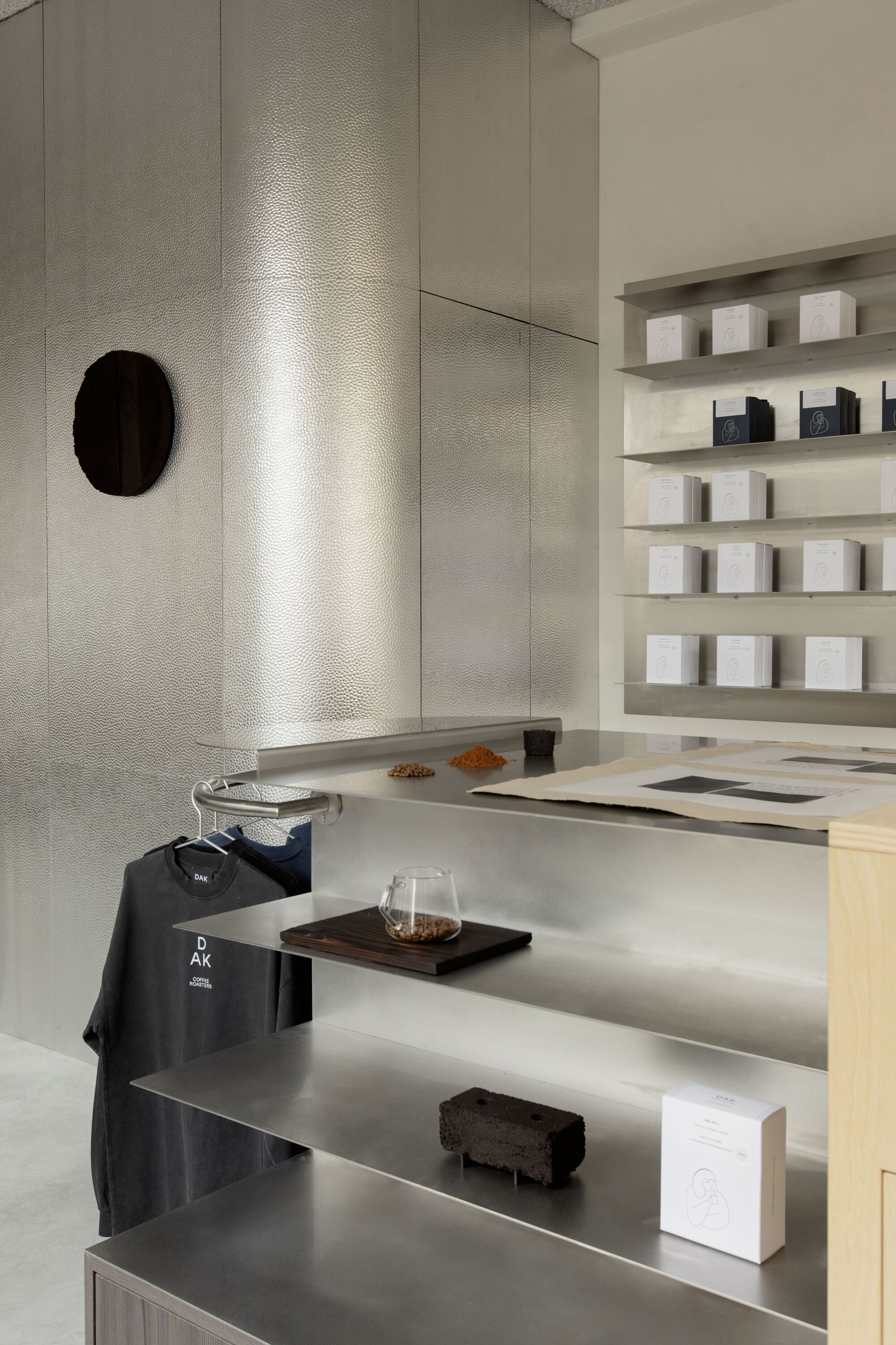 Hammered steel wall and folded metal display shelves holding boxed coffee, a glass server and a hanging T-shirt