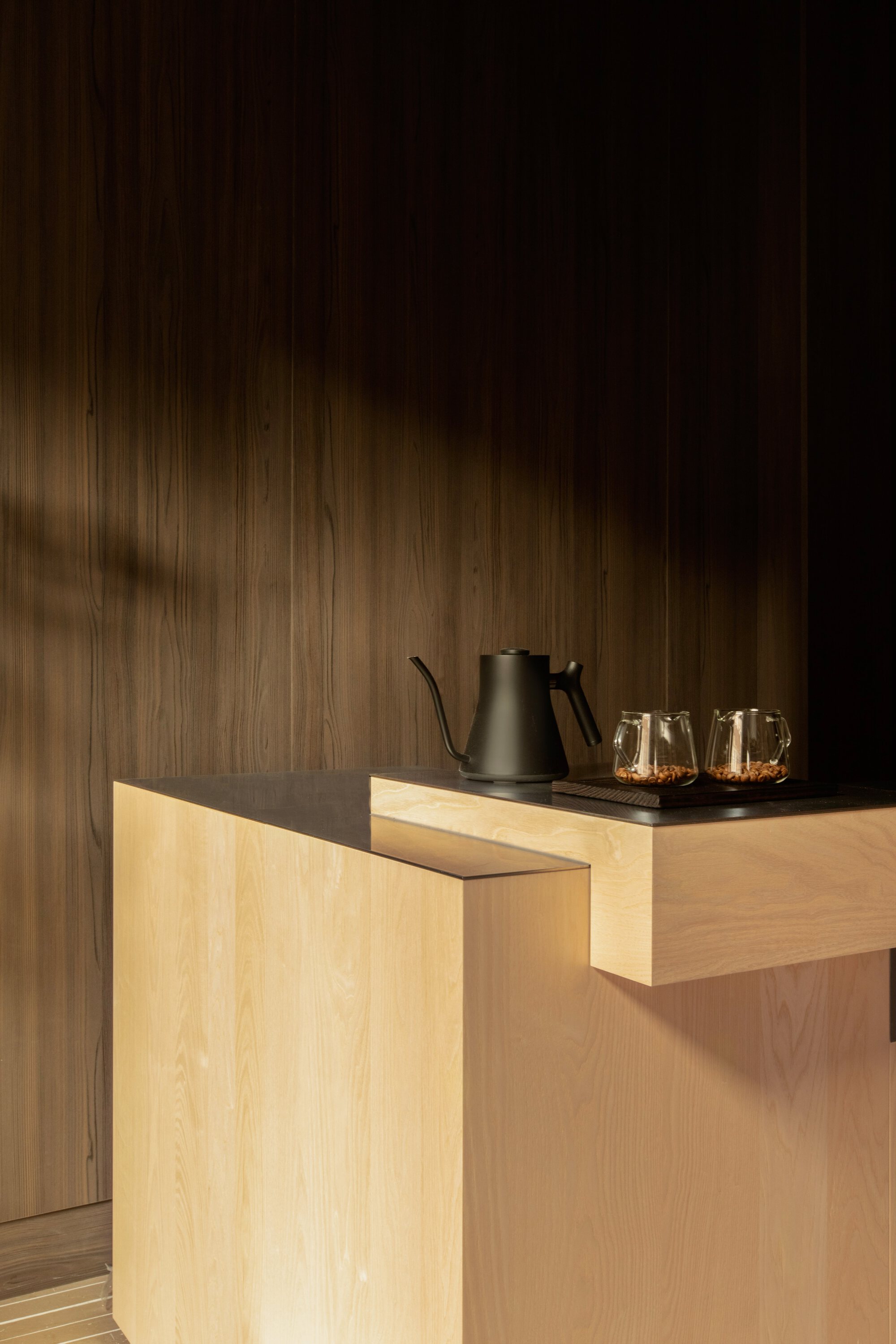 Oak counter corner with a black kettle and two glass servers against dark wood panelling, with a soft shadow across the wall