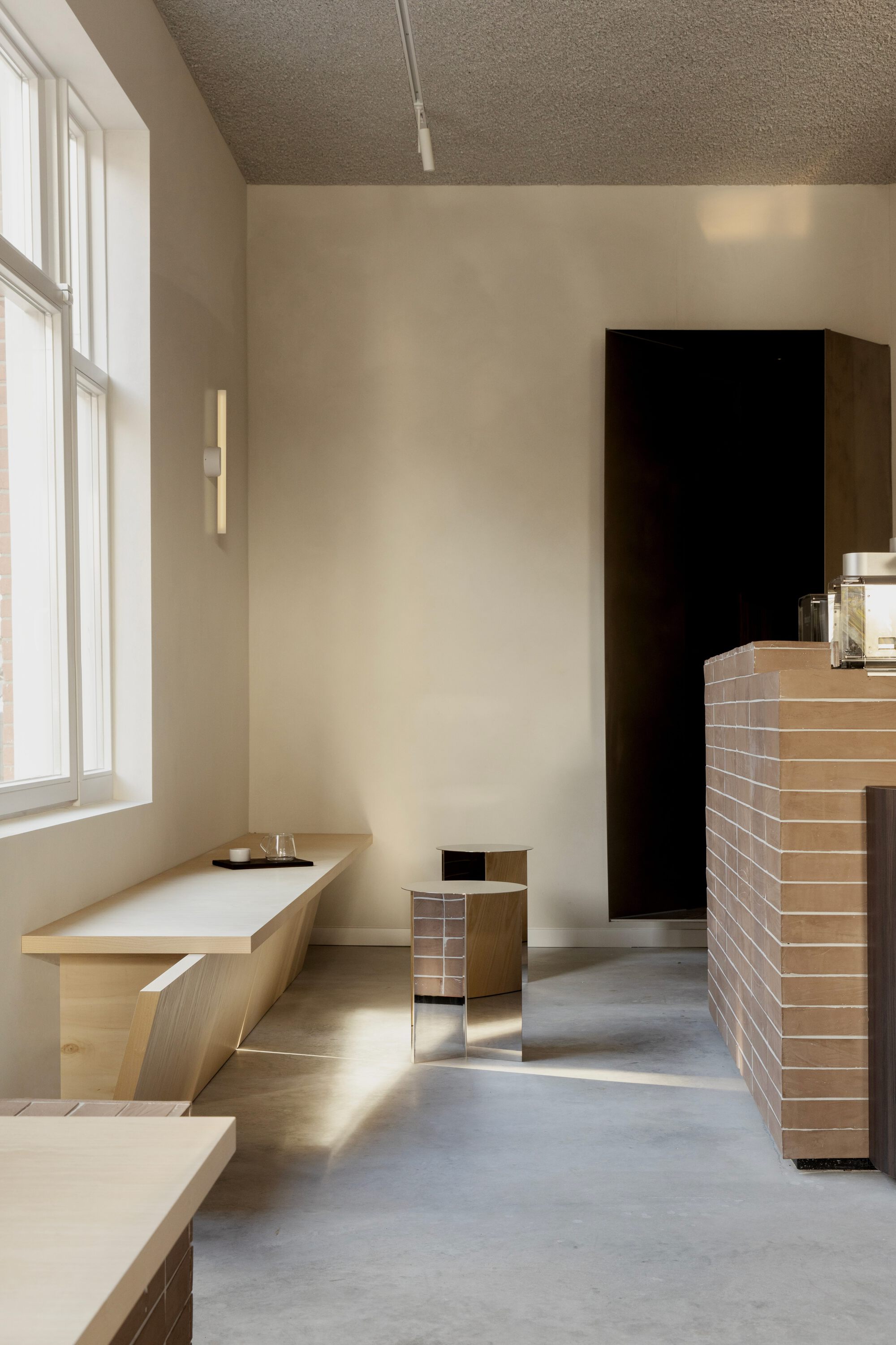 Pale timber bench and mirrored stools on a concrete floor, with sunlight falling through a tall window beside the brick counter