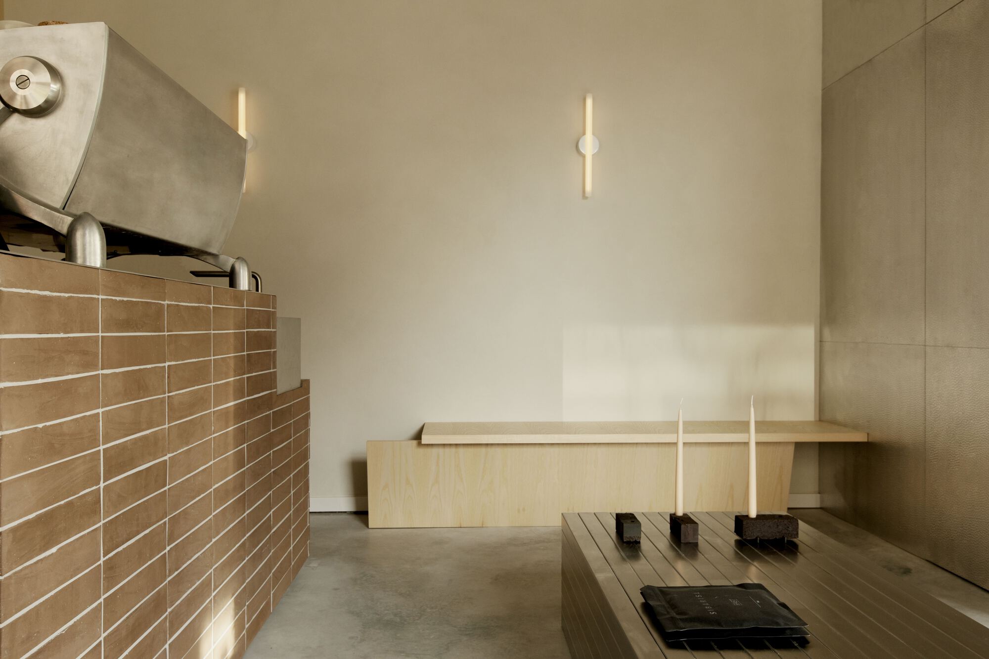 Brick-clad counter with a steel coffee roaster beside a pale timber bench and slatted table, lit by soft window light