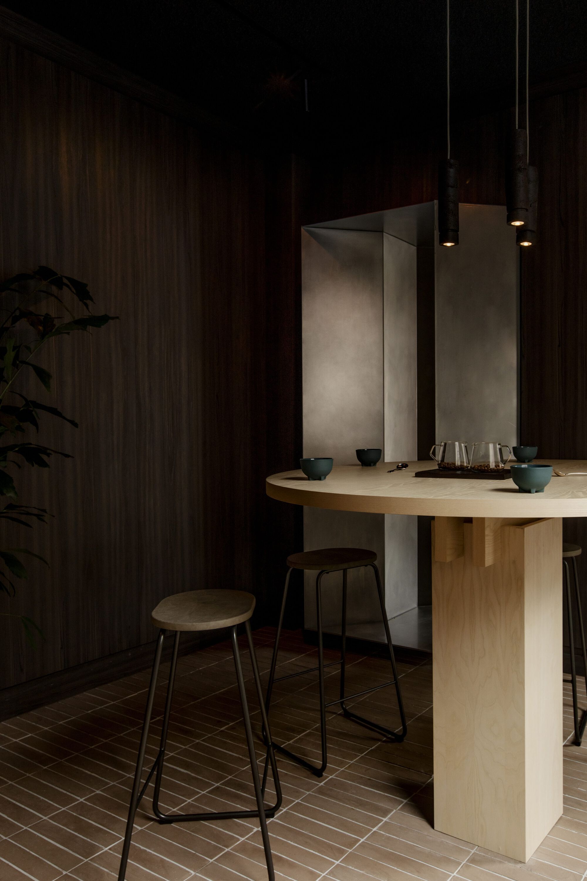 Round pale timber table with teaware and stools in front of a steel alcove, under pendant lights in a dark room