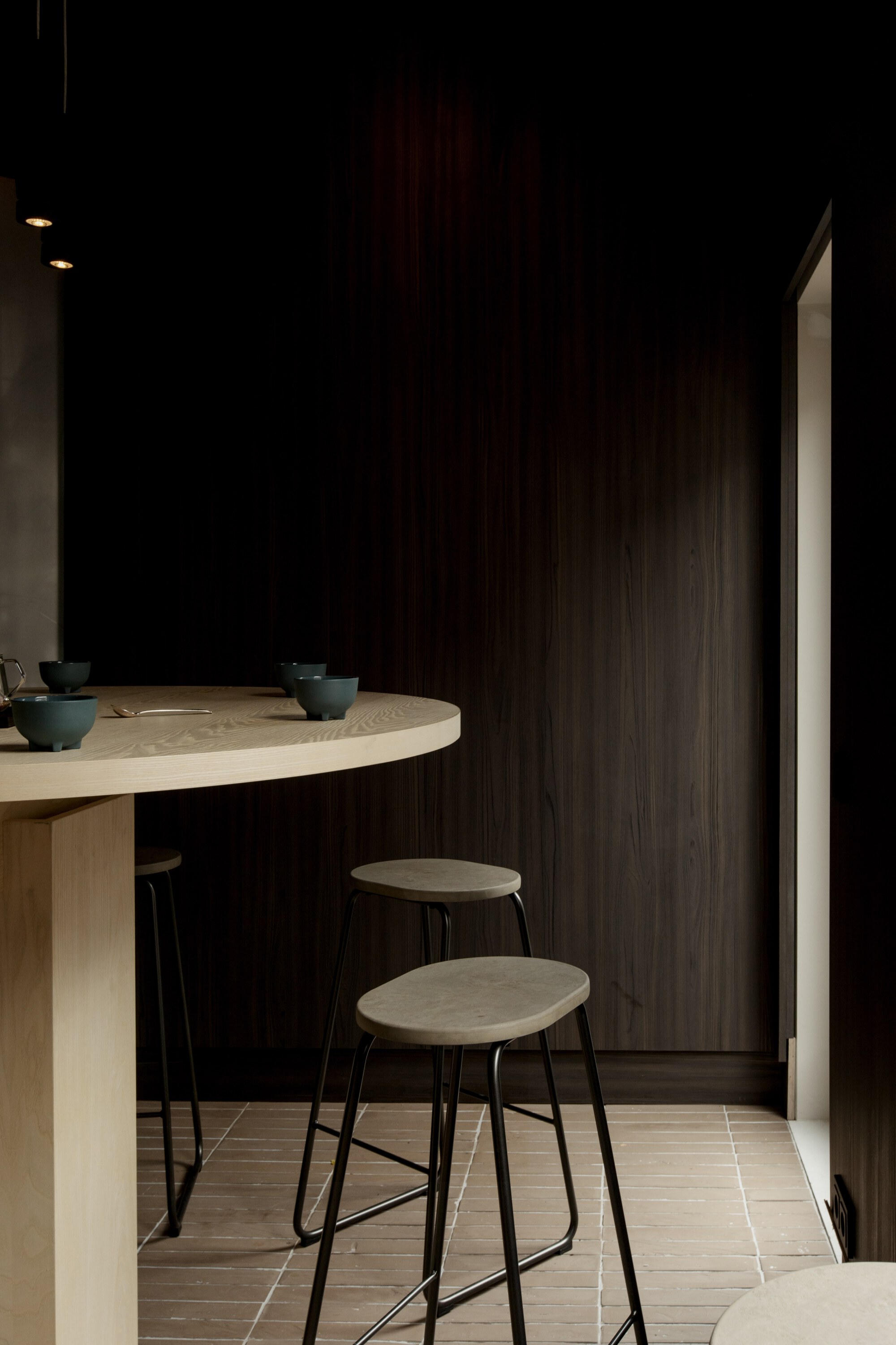 Black-framed stools beside the round table edge, set against dark wood paneling with daylight at a narrow window