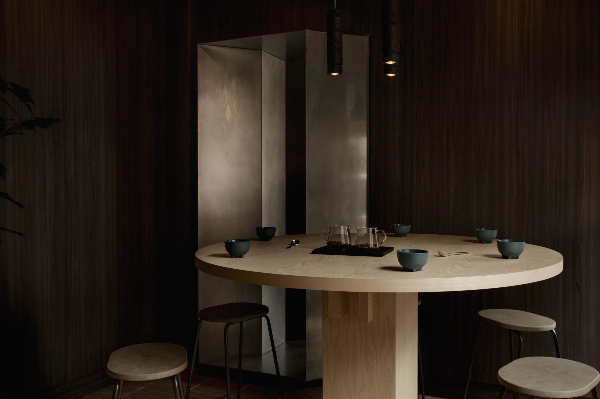 Round ash table set with green bowls and glass carafes before a steel niche, lit by low pendant lamps