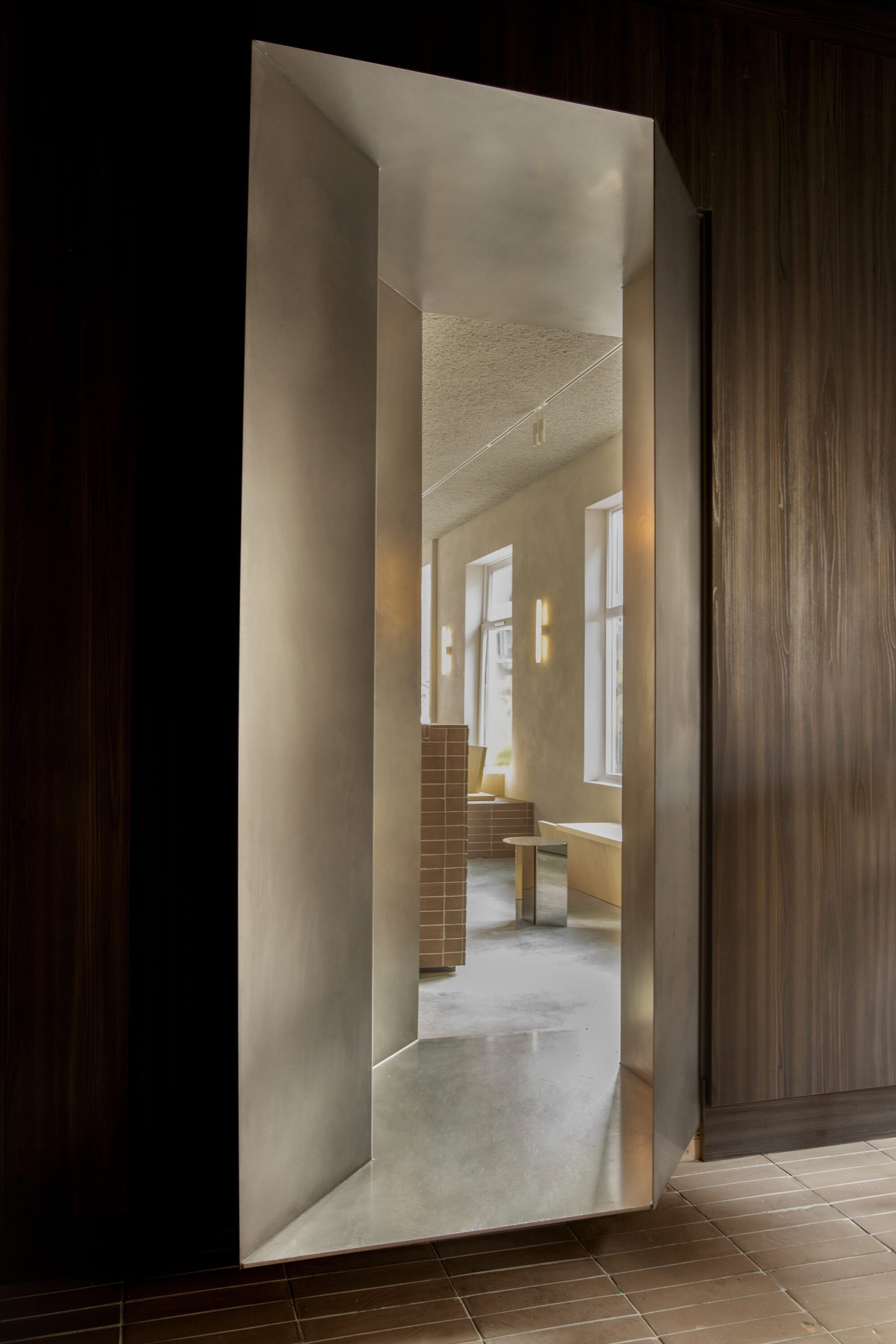 Brushed metal framed opening in a dark wood wall, revealing a tiled counter and windows in the room beyond