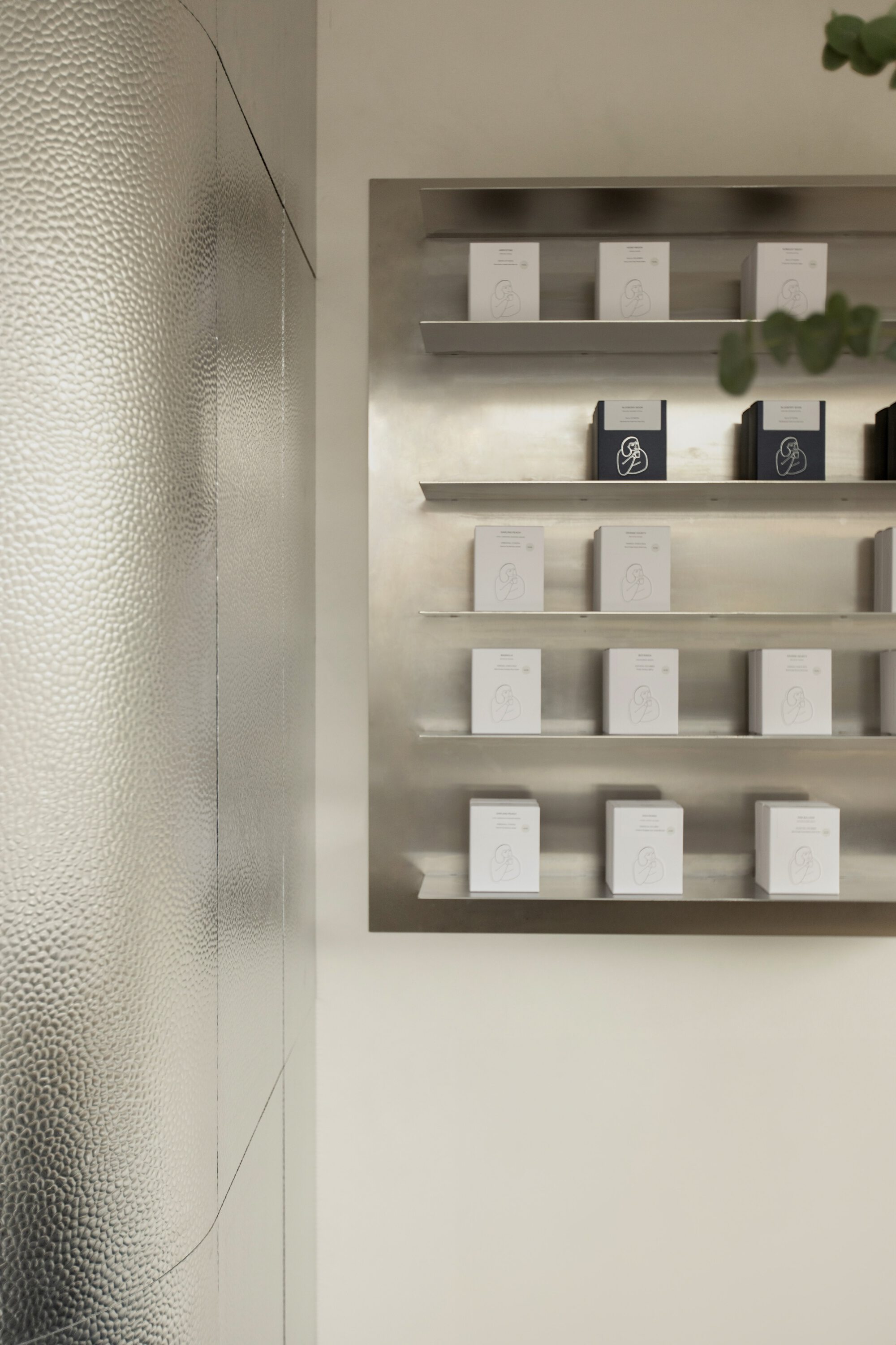 Steel wall shelf holding rows of white and navy boxed candles beside a hammered metal curved wall panel