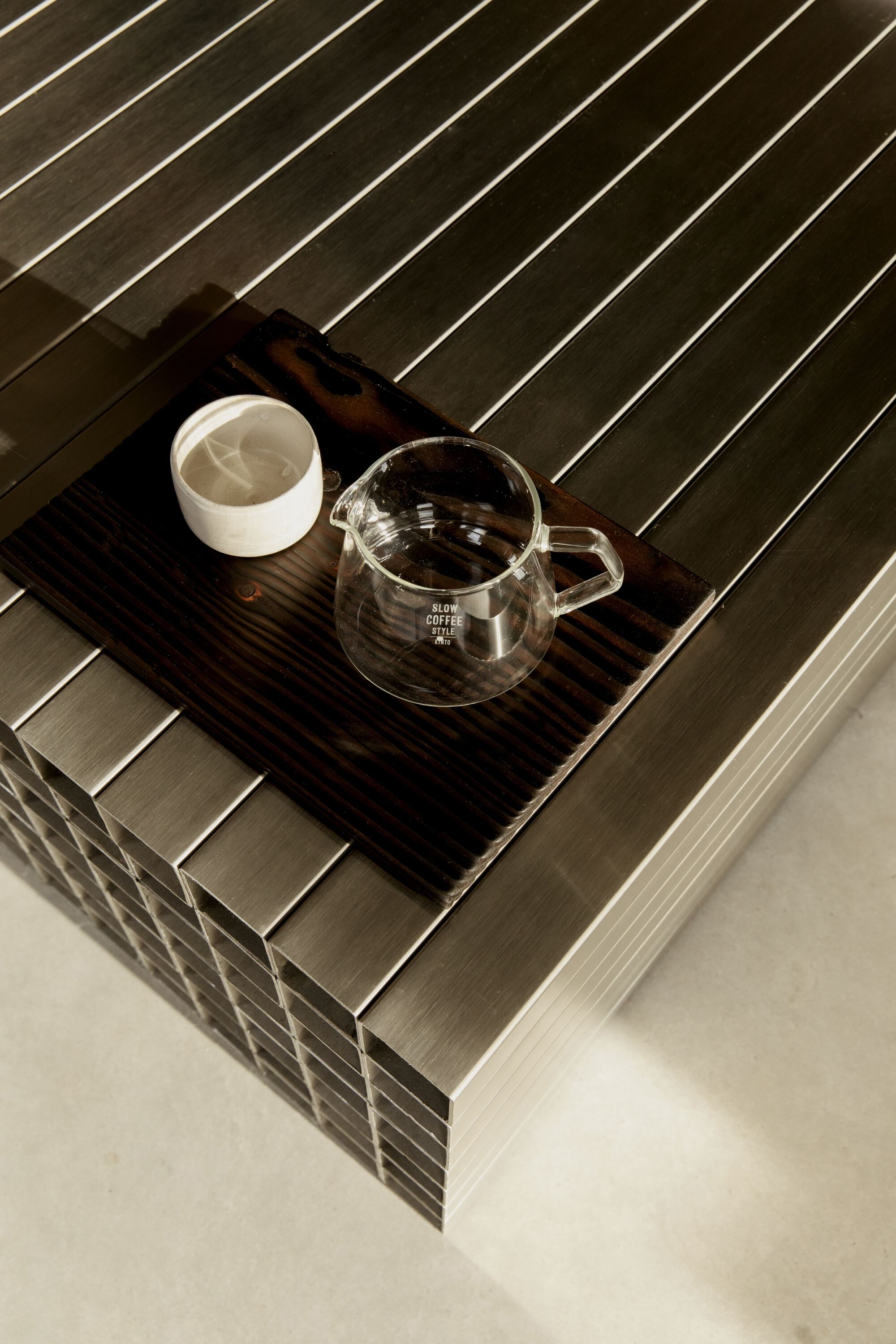 Overhead view of a glass coffee server and cup on a charred wood tray atop a slatted steel counter