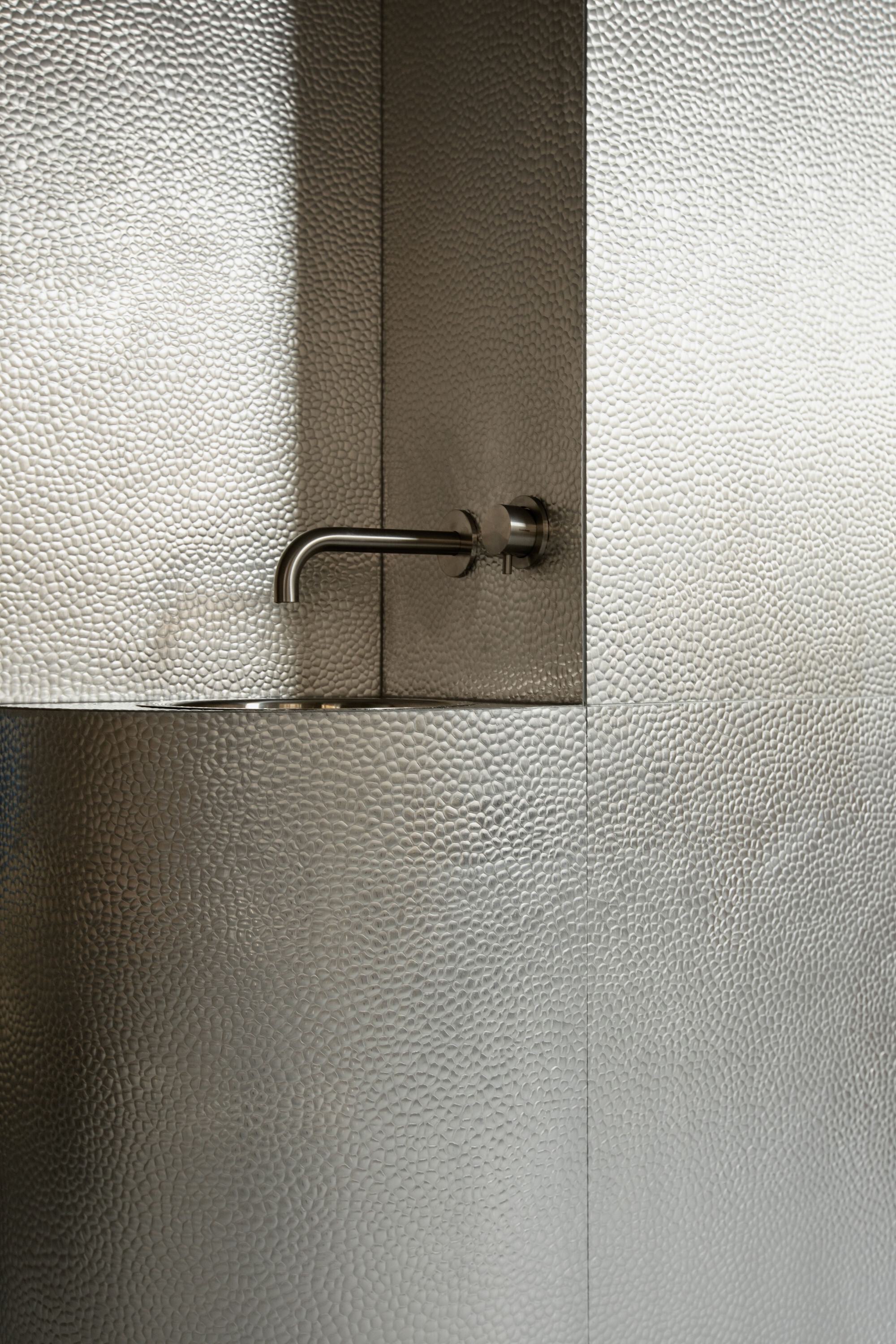 Wall-mounted brass tap and basin set into hammered metal panels, with soft light falling across the surface