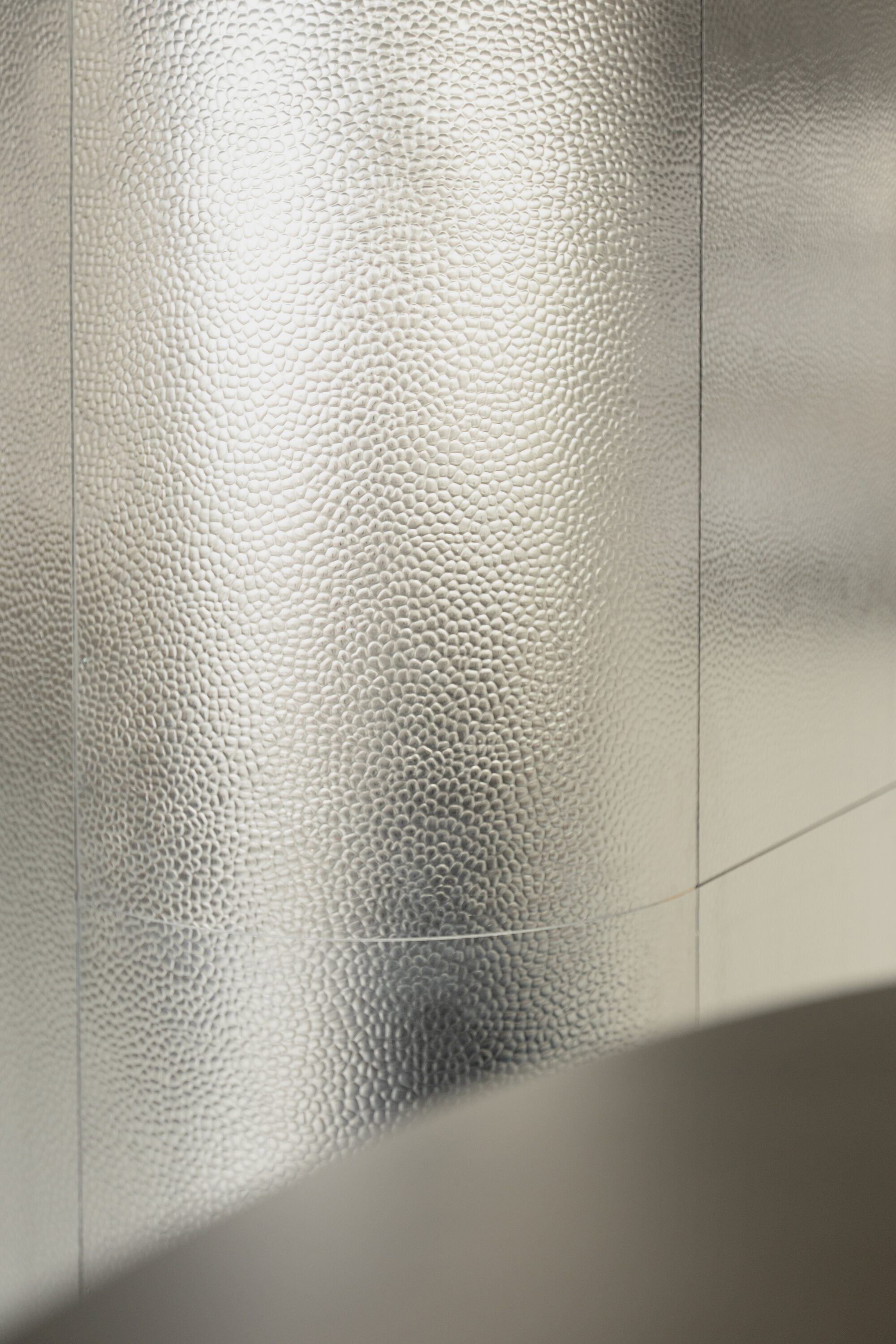 Close-up of a curved hammered metal wall panel with fine seams, catching diffuse window light above a basin edge