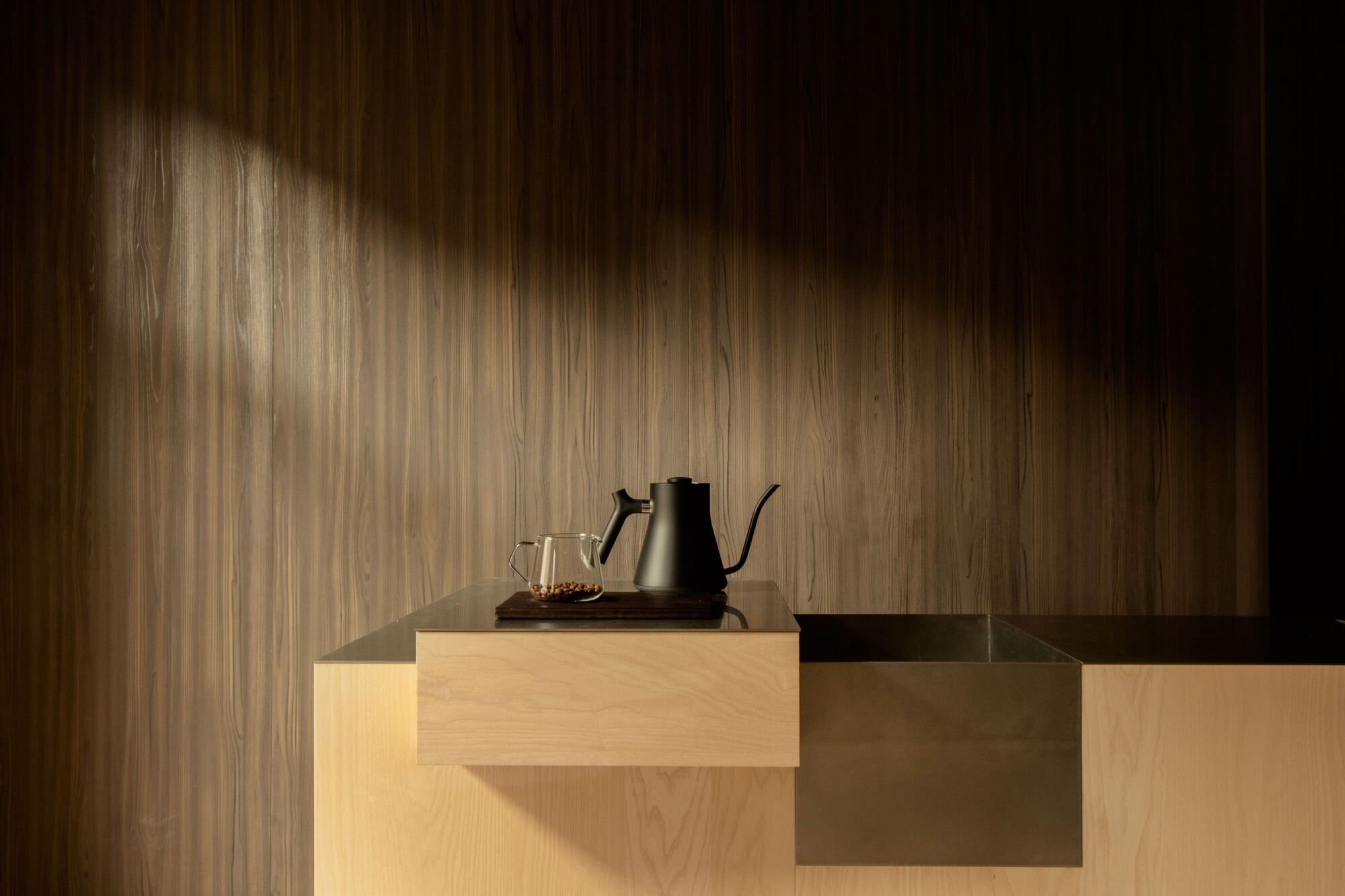 Black kettle and glass server on a pale timber counter against a walnut wall, crossed by a beam of sunlight