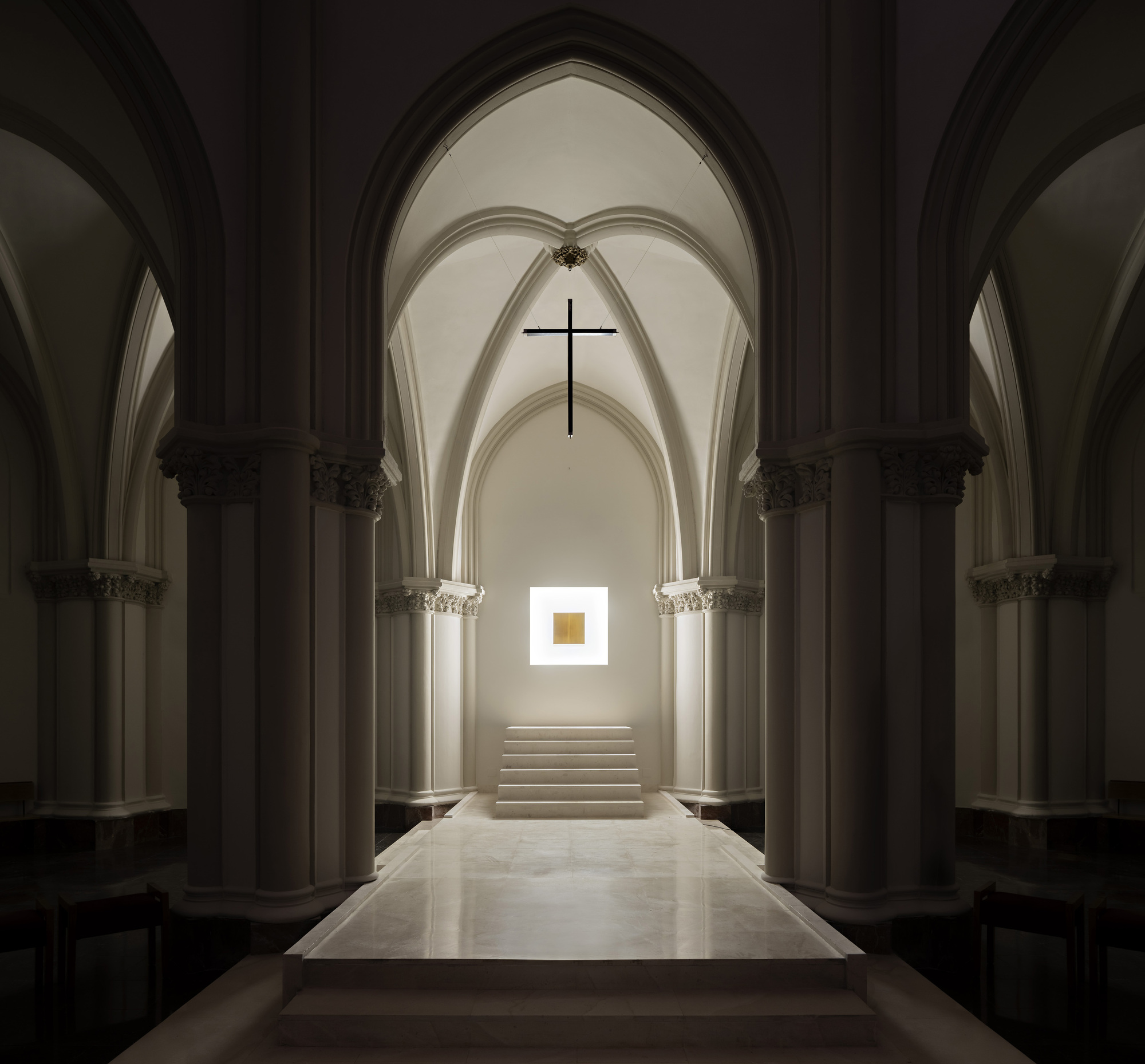 Central nave axis of a Gothic chapel with a black cross above a stepped marble platform and a glowing square niche in dim light