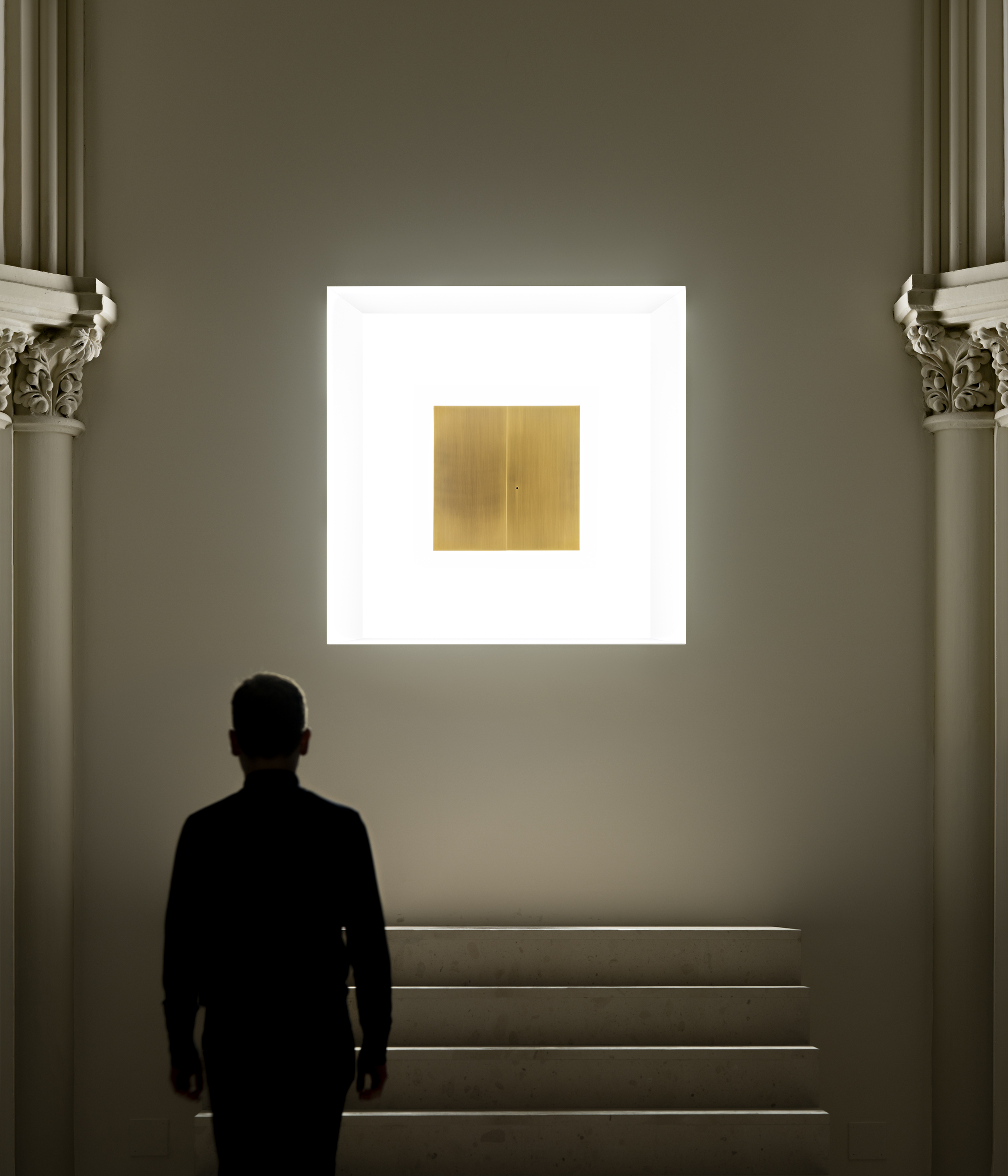 A silhouetted visitor faces a backlit square niche holding a brass panel above a short flight of stone steps