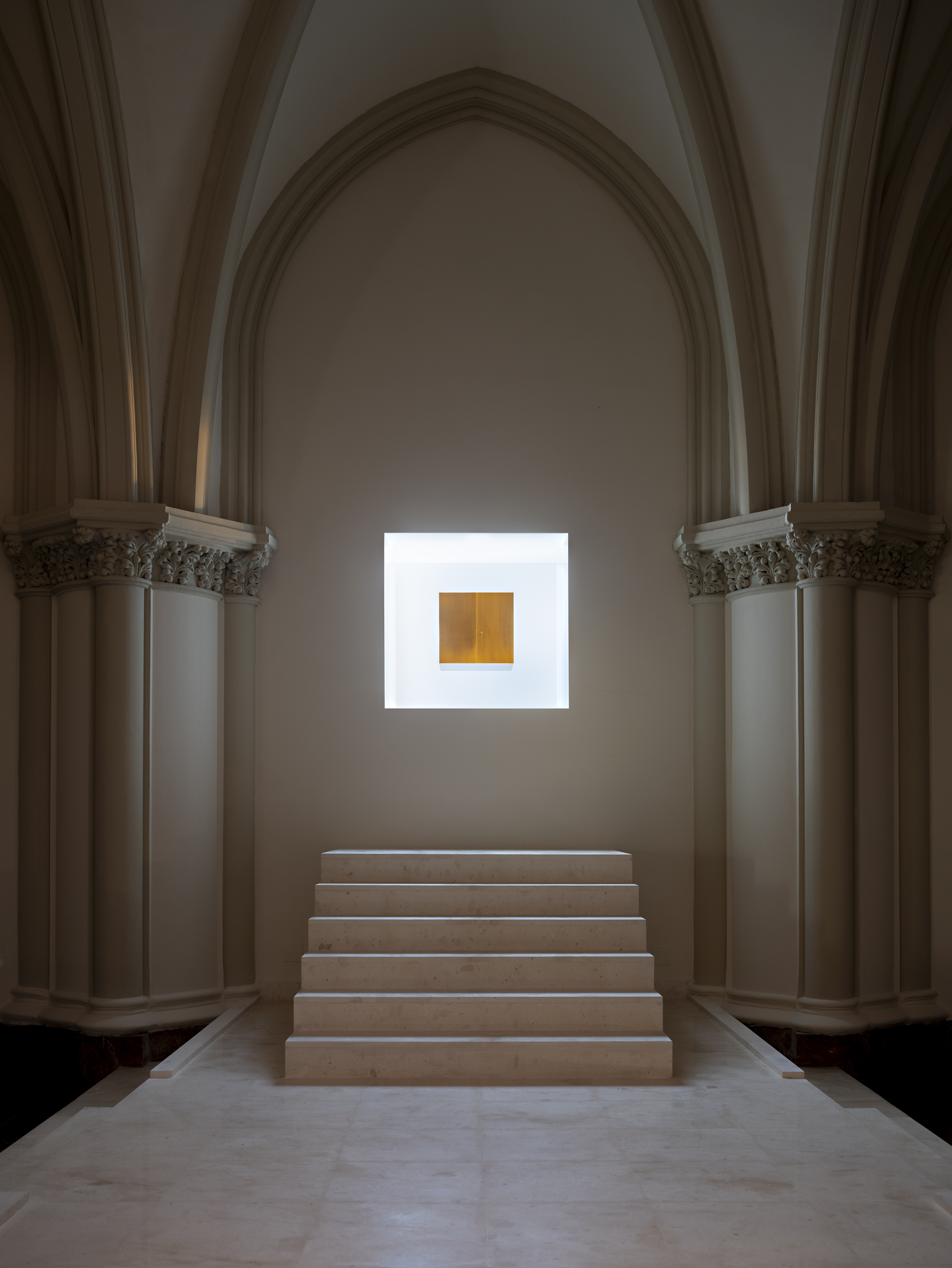 Symmetrical view of five stone steps rising to a luminous square niche with a brass panel between carved column capitals
