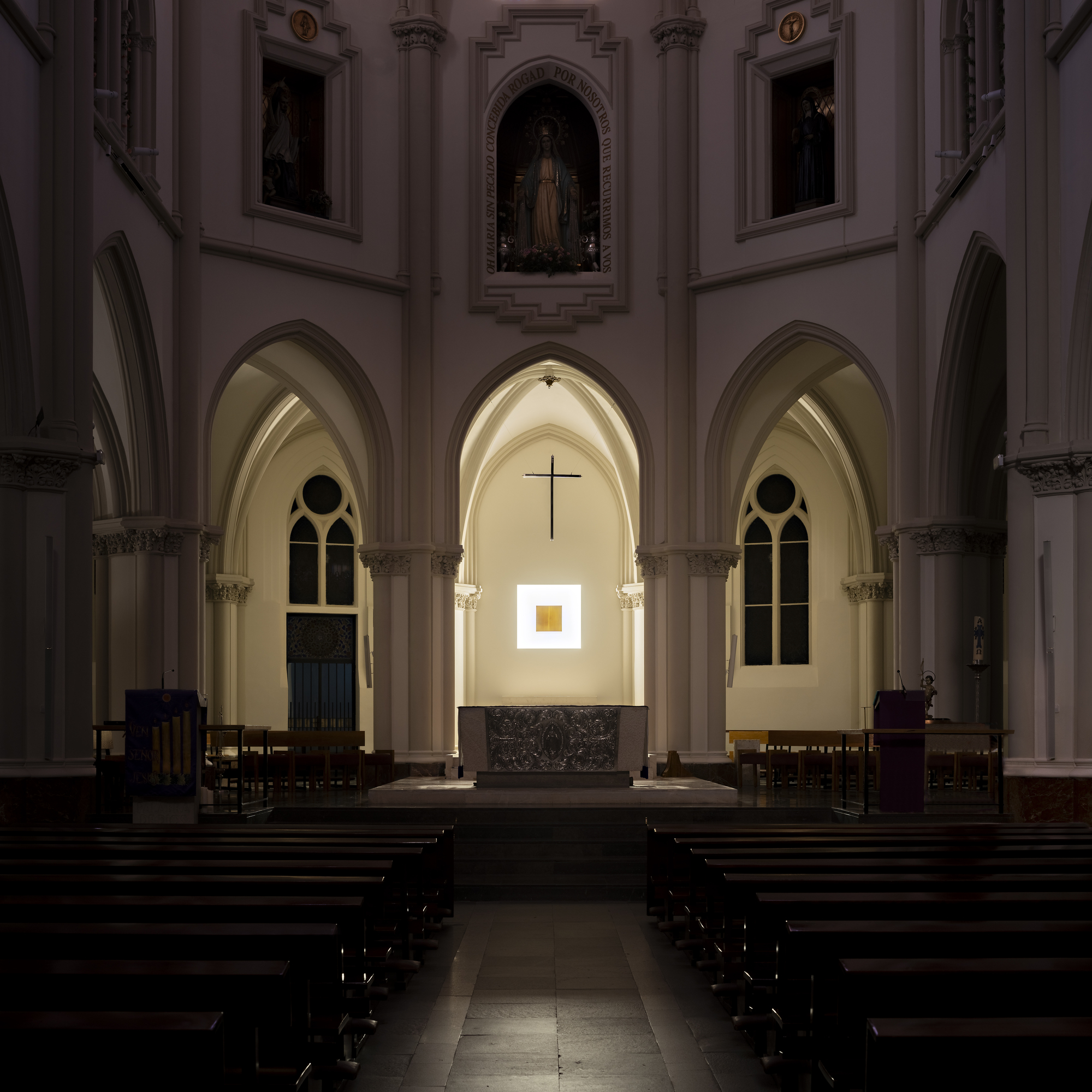 Long view down the church nave past dark pews to the altar, with a cross and glowing square niche in the apse