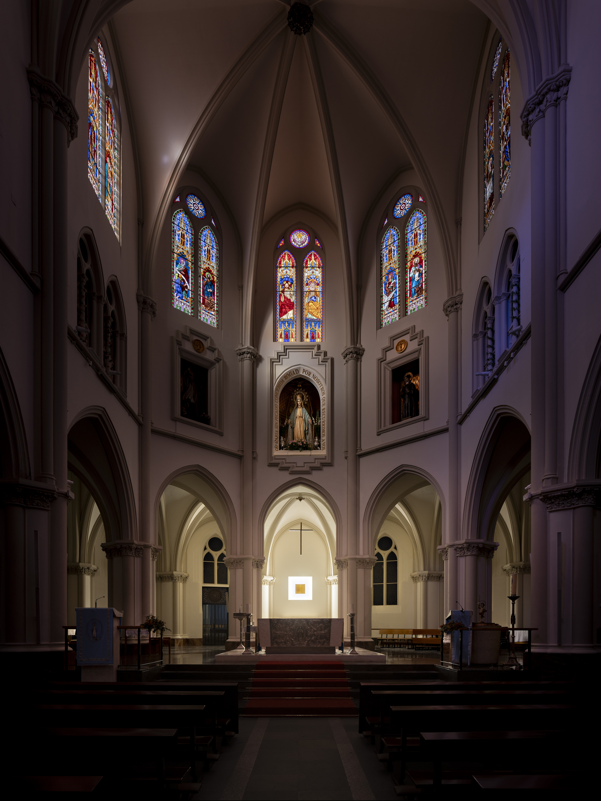 Nave view toward the apse, with stained glass windows and a small lit panel above the altar in a darkened church