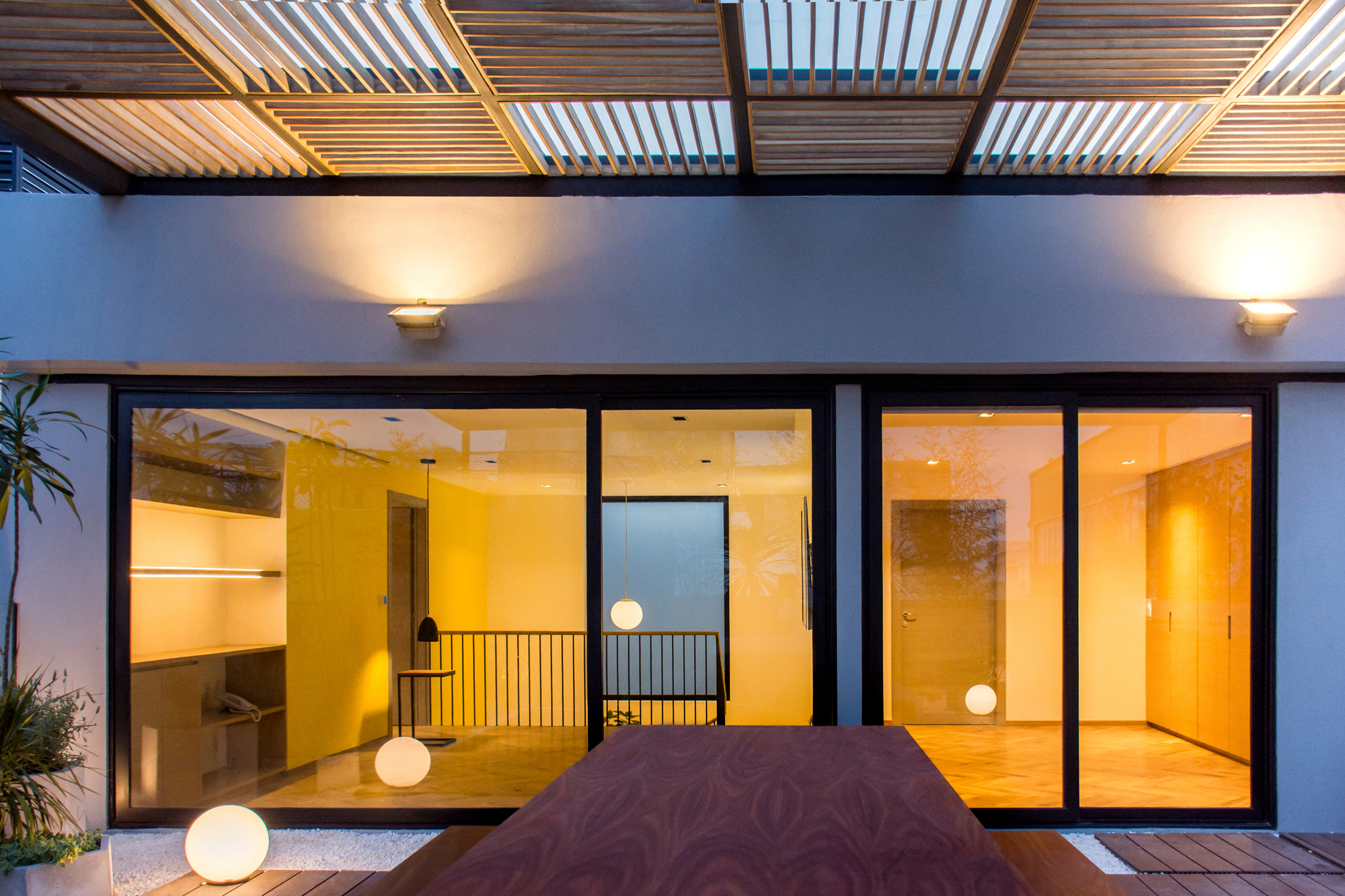 Terrace dining table in front of glazed sliding doors, with a slatted timber pergola overhead and globe lamps glowing at dusk