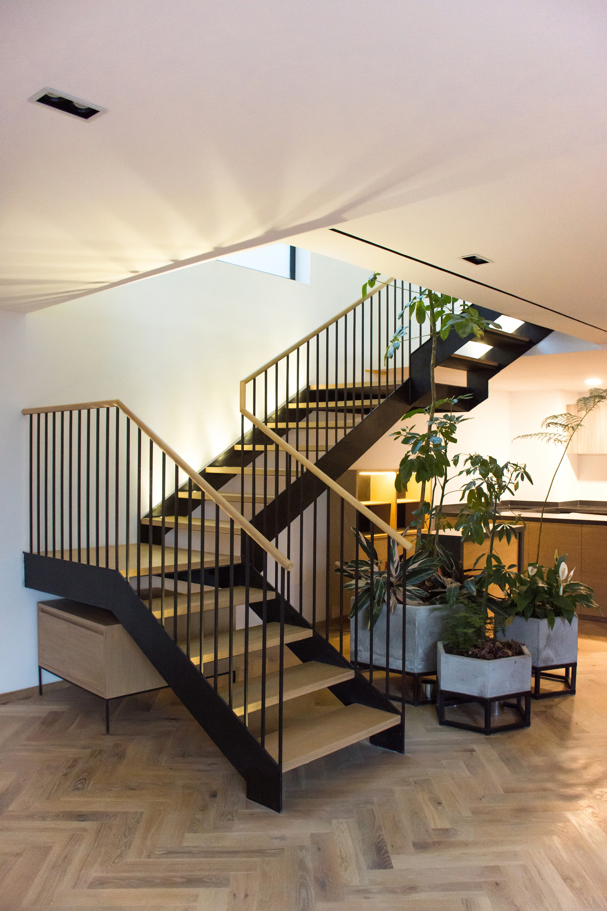 Black steel stair with timber treads and vertical balusters rising above a herringbone floor, with potted plants in concrete planters beside it