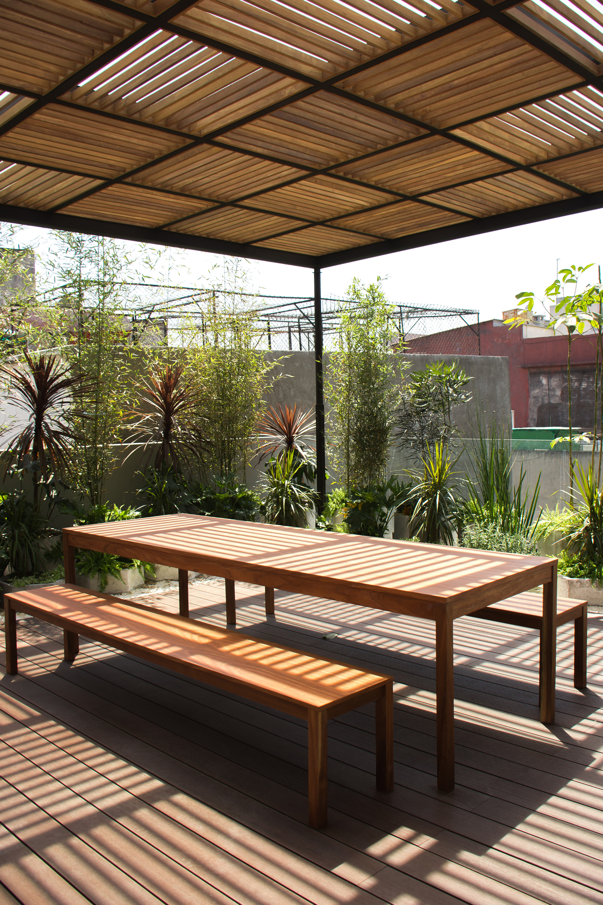 Timber table and benches on a deck under a slatted pergola, casting striped sunlight shadows in front of bamboo and grasses