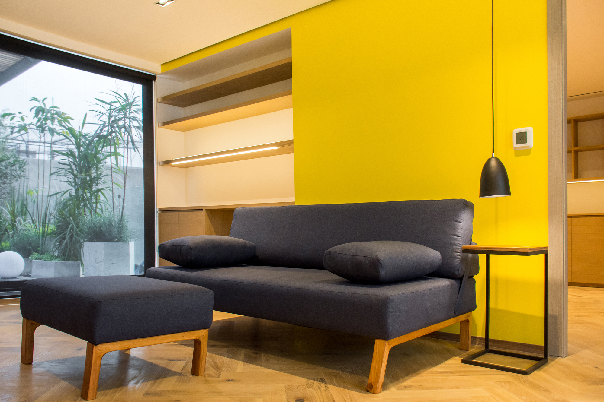 Grey sofa and ottoman with timber legs against a yellow wall with built-in shelves, a black pendant lamp hanging beside them