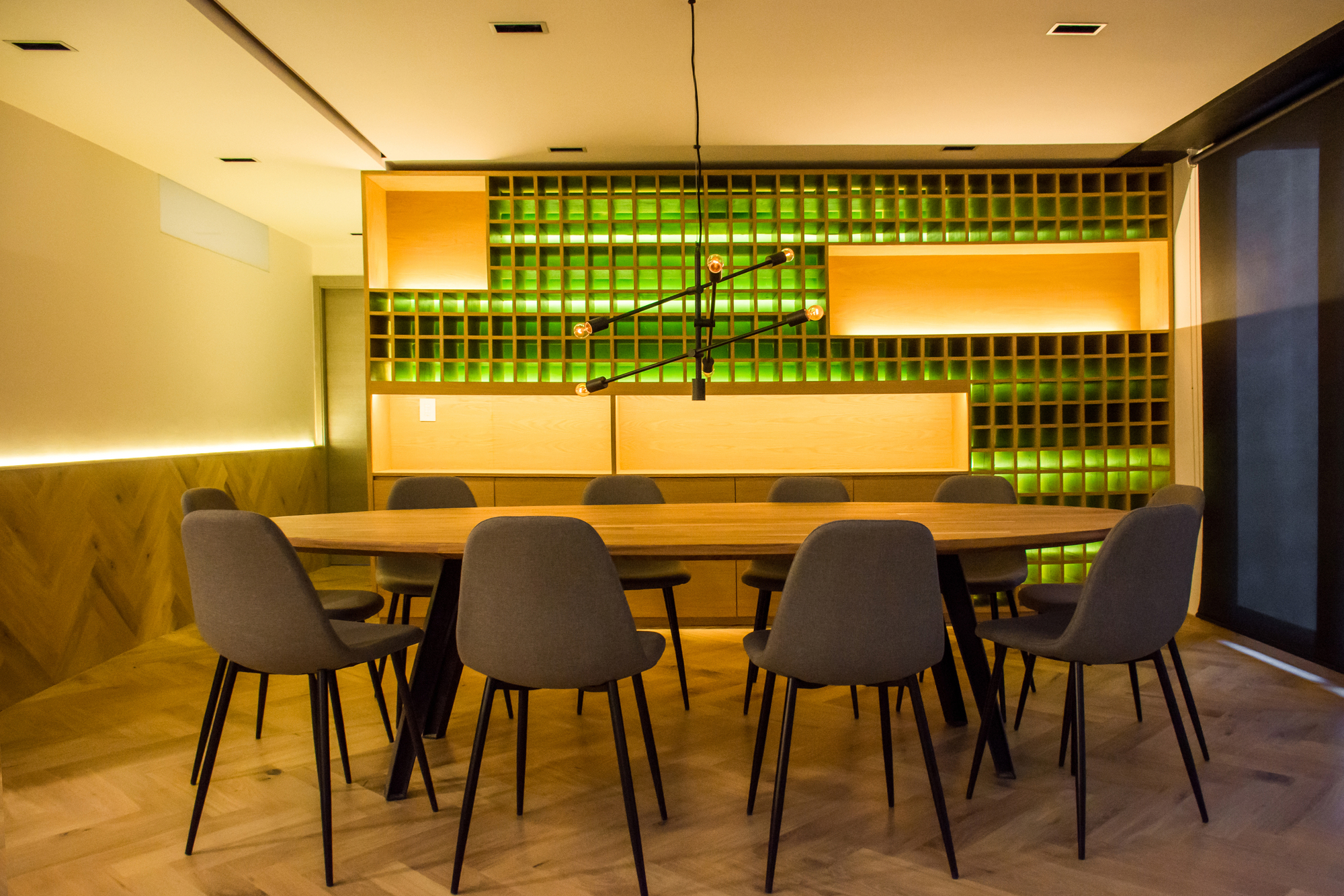 Oval dining table with grey chairs facing a gridded shelving wall lit green, with a branching pendant light above in the evening