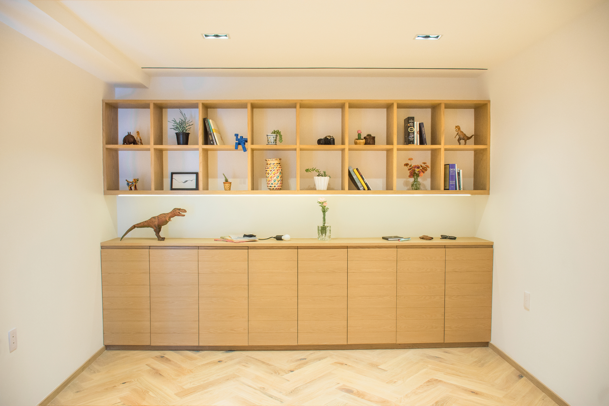 Oak wall of open cubby shelves above flush base cabinets, with toy dinosaurs and potted plants under a soft strip light