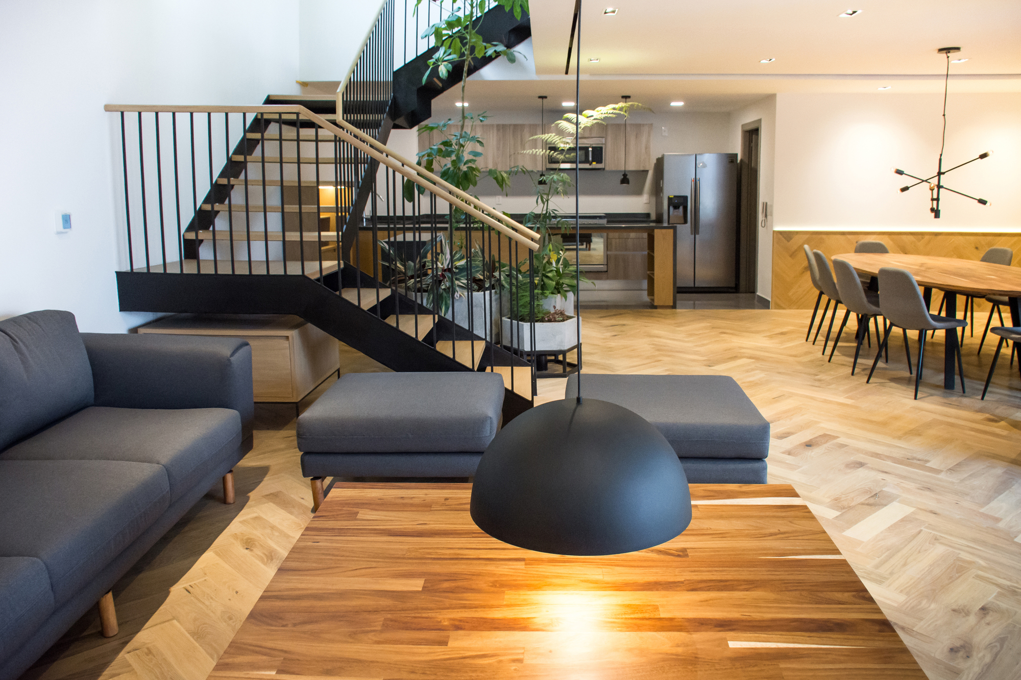 Living area with a black steel stair, grey sofa and ottomans, and a pendant lamp over a walnut table in warm light