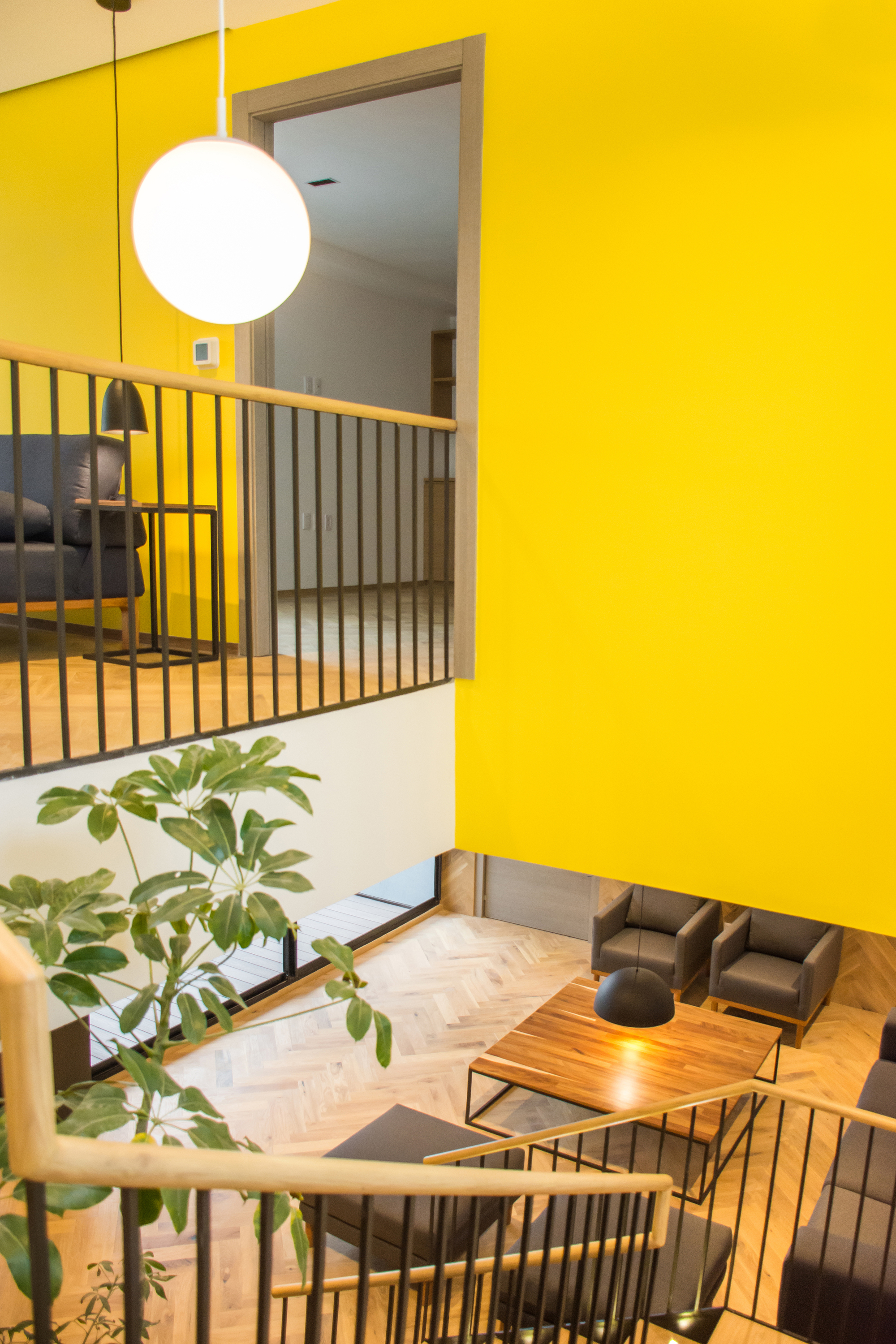 View down from the upper landing past a steel baluster rail and leafy plant to the seating area below a yellow wall
