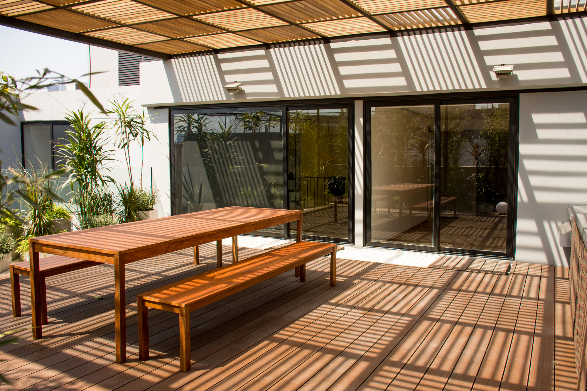 Roof terrace with a timber table and benches on decking, striped by shadows from the slatted pergola in afternoon sun