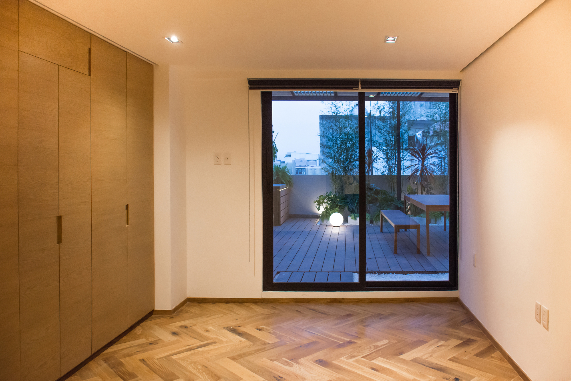 Bedroom with full-height oak wardrobe doors and a glass door opening to the terrace, where a globe light glows at dusk