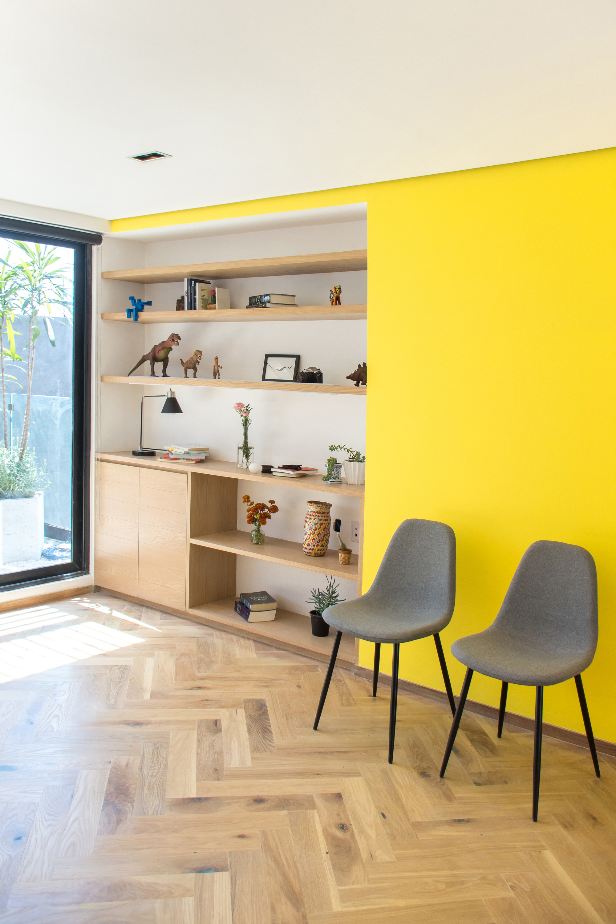 Built-in oak shelving beside a yellow wall, with two grey chairs on herringbone flooring in bright window light