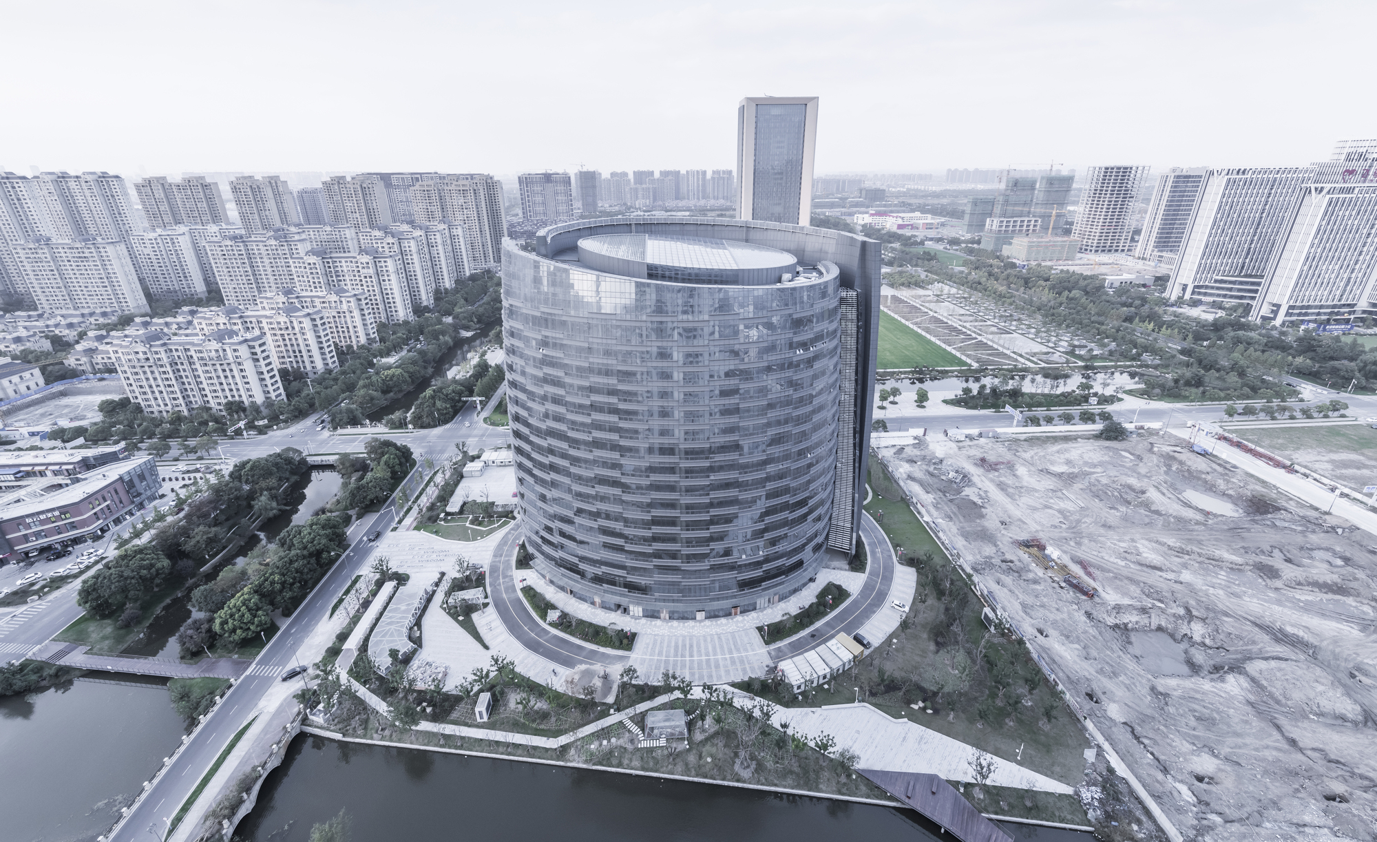 Aerial view of the oval glass tower ringed by a curving drive, canal and cleared construction land under hazy sky