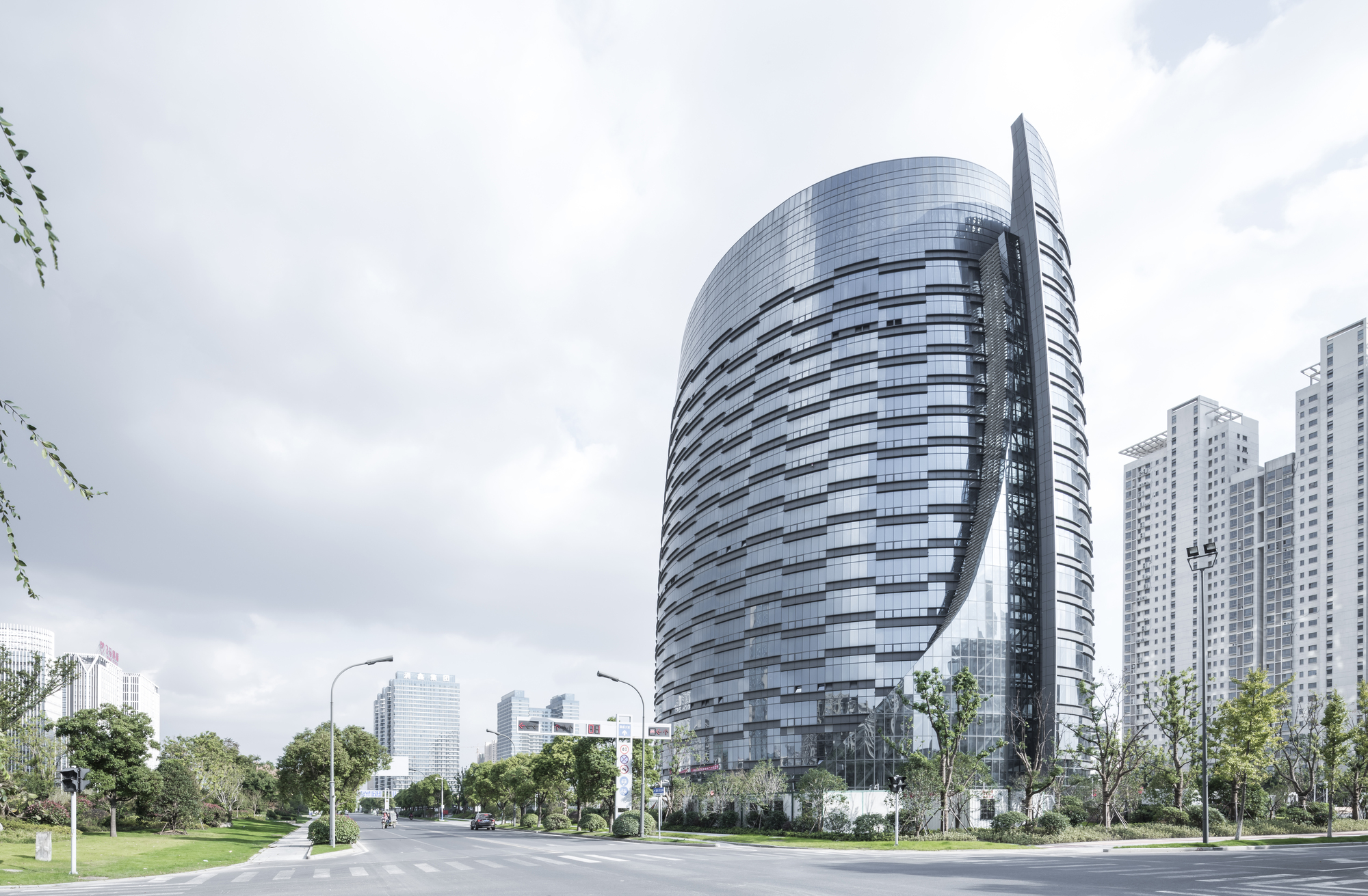 Street-level view of the curved glass tower with its vertical steel fin, set against overcast sky and roadside trees