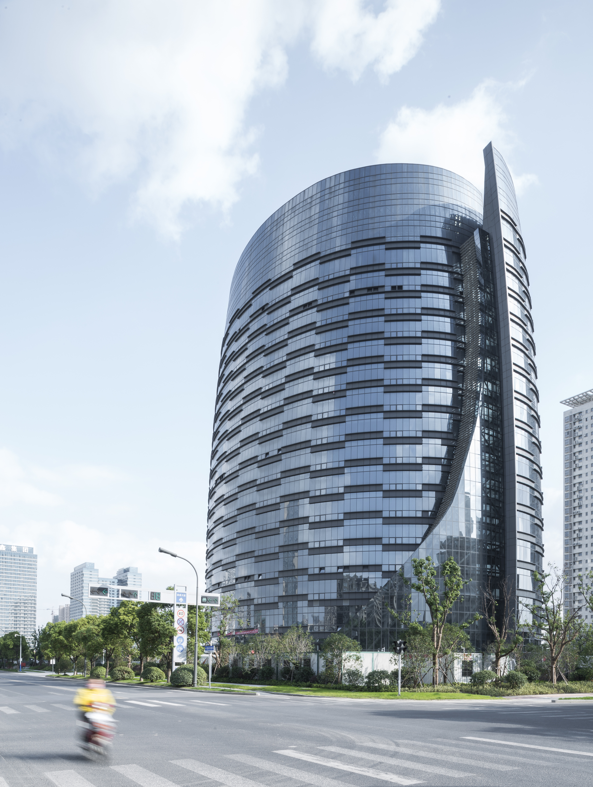 Tall glass tower with horizontal banding and a slim vertical blade, a scooter blurring past on the crossing below