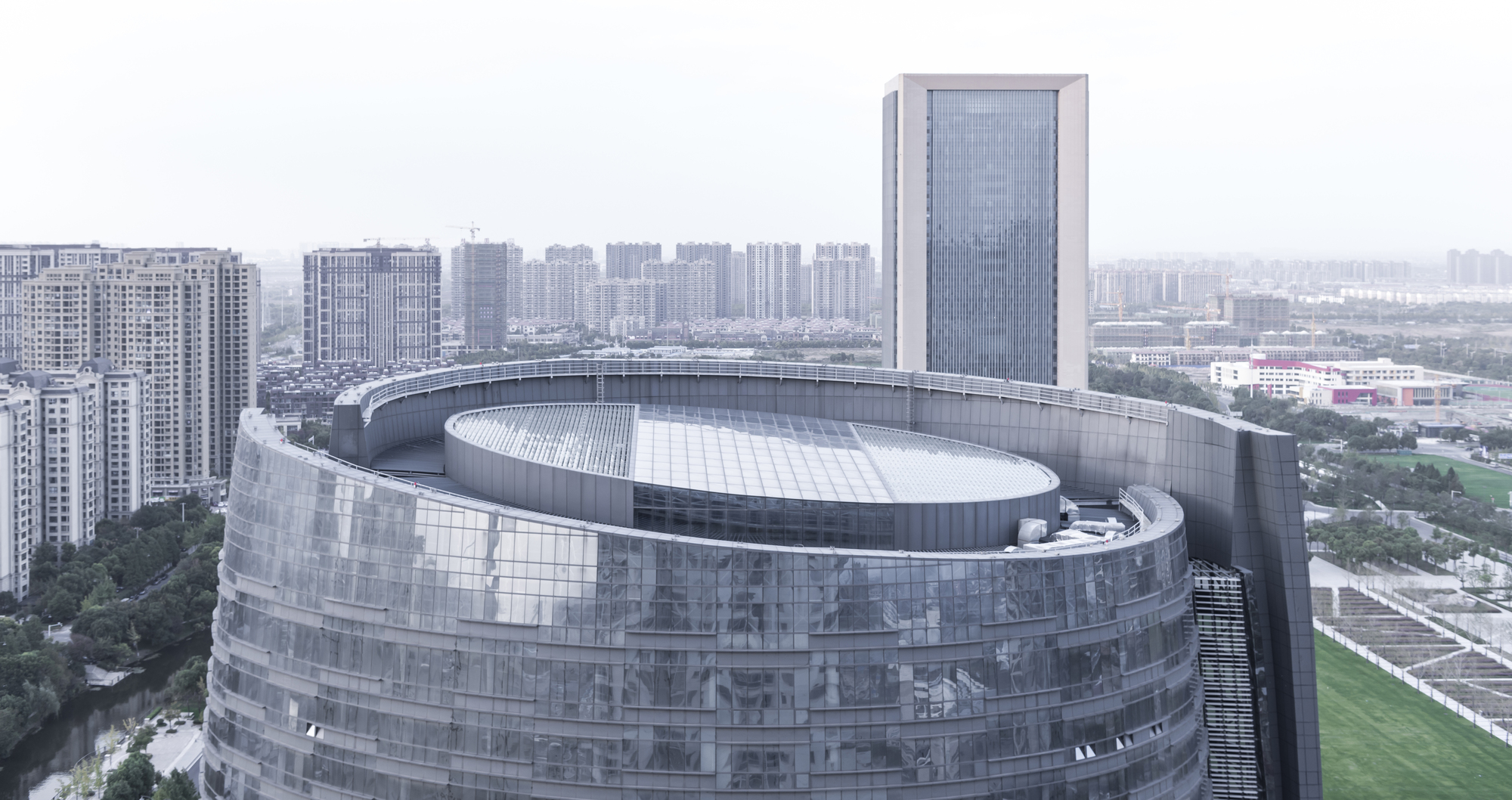 Rooftop view of the stepped oval crown with a glazed skylight disc, green sports field and apartment blocks in haze