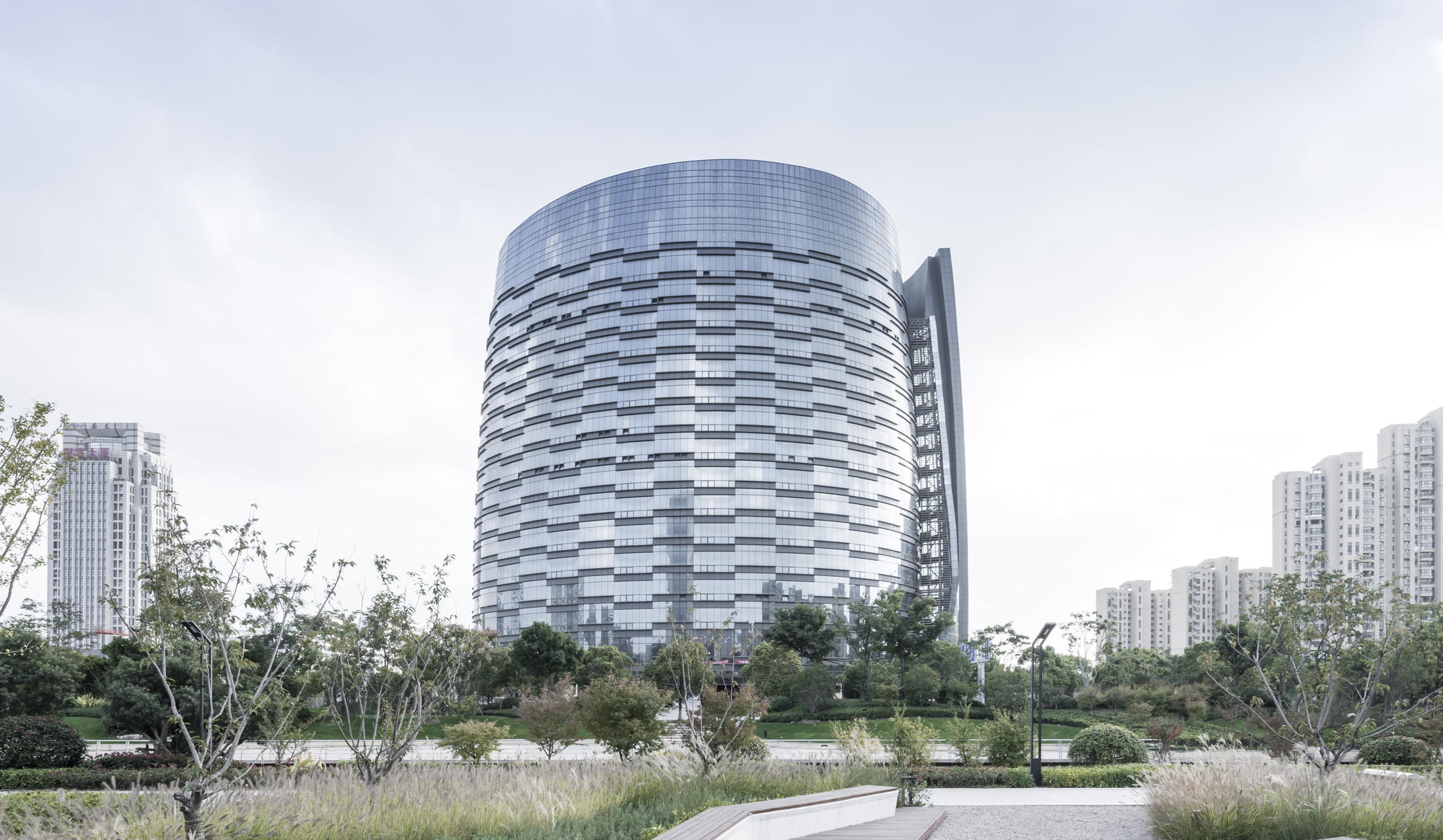 Cylindrical glass tower with a slim fin tower at its side, seen across a planted park under overcast sky