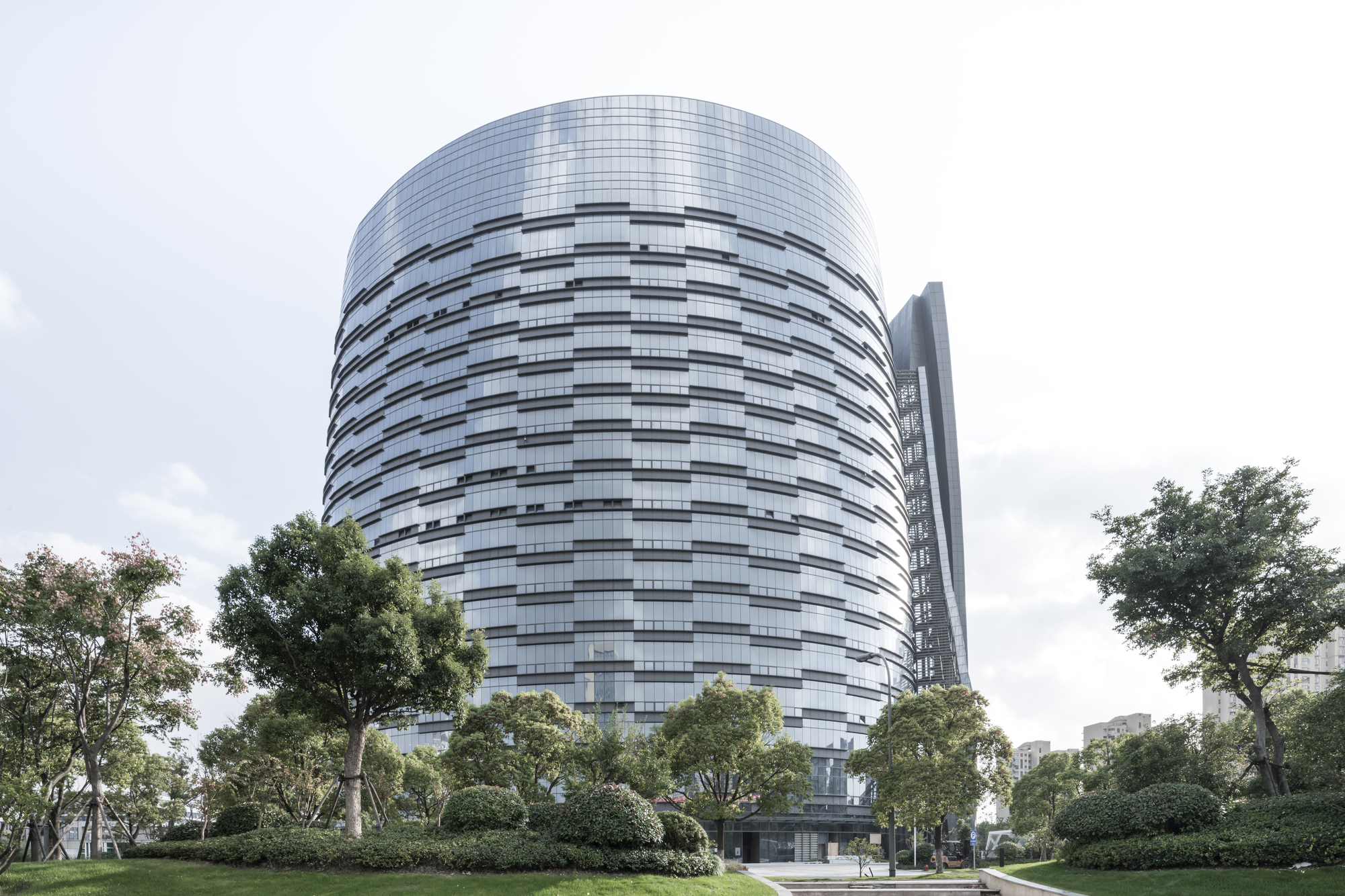 Low view of the banded glass tower rising behind clipped shrubs and trees under a pale cloudy sky