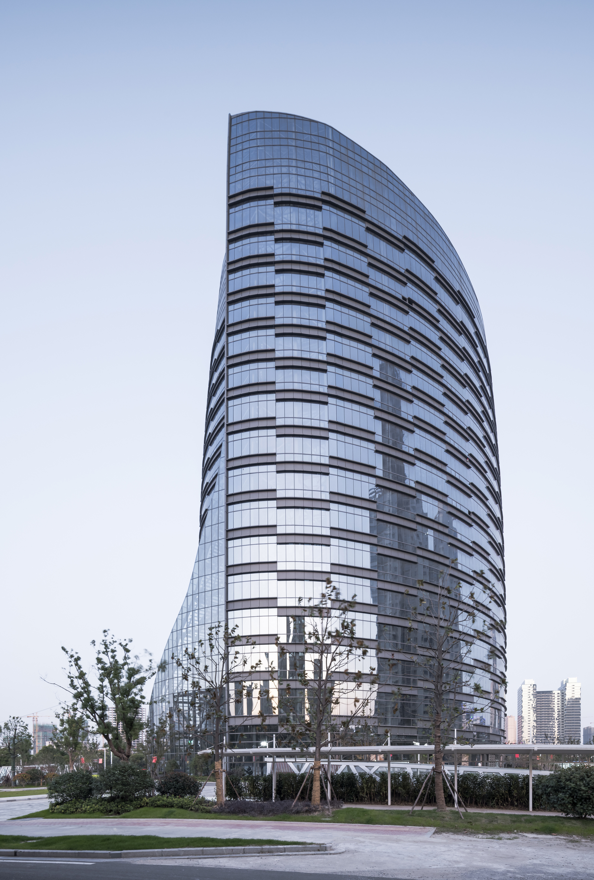 Tapered glass tower with a slanted curved top, framed by young staked trees in clear dusk light