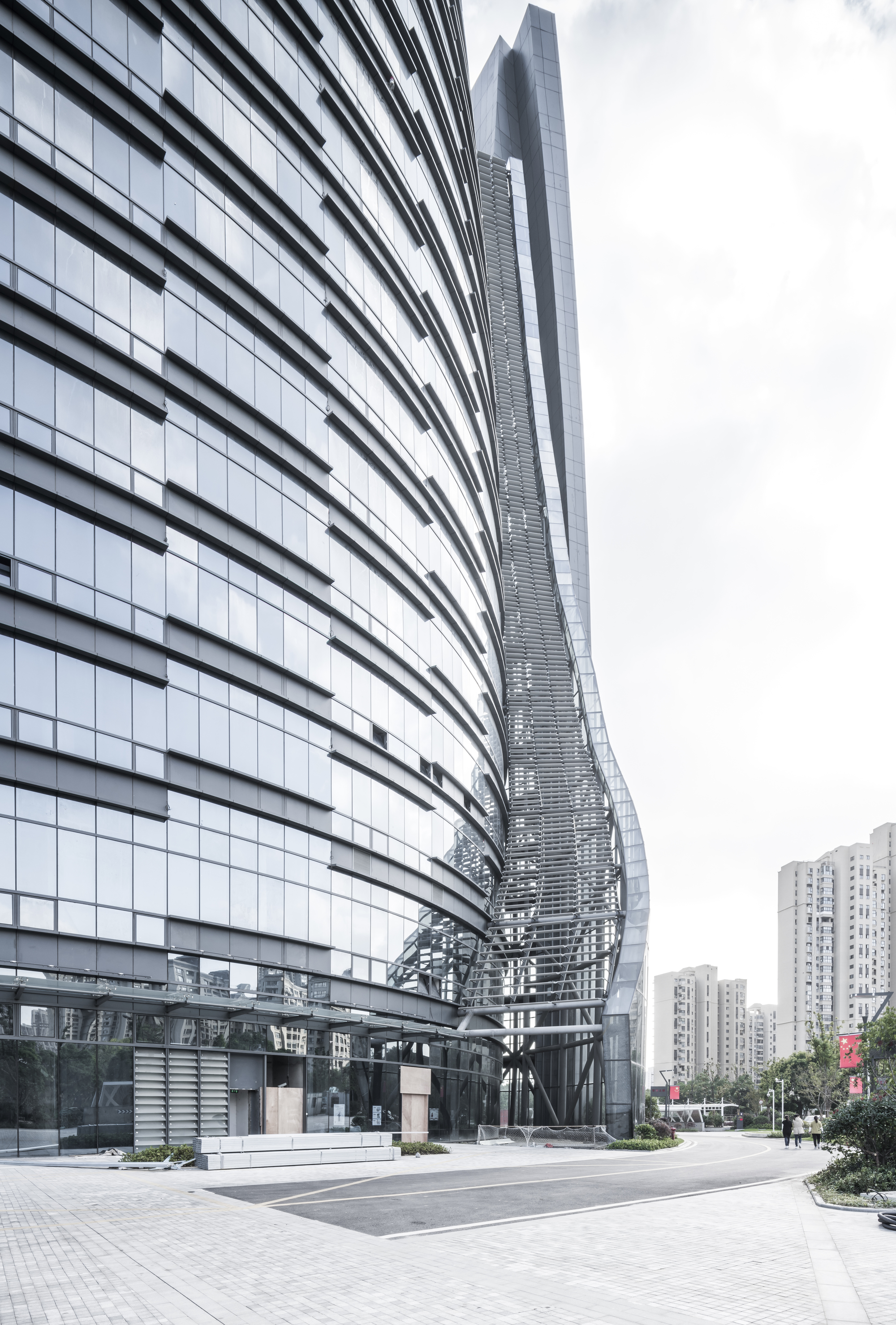 Base of the tower where the curved glass wall meets a latticed vertical spine, with pedestrians on the paved plaza
